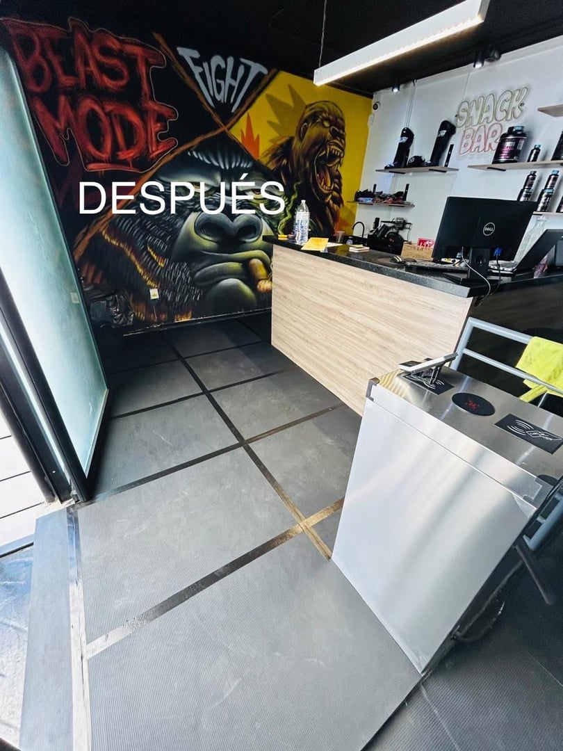 Gym reception area with counter, mural of gorilla and fighter, labeled DESPUÉS, featuring a turnstile and snack bar sign.