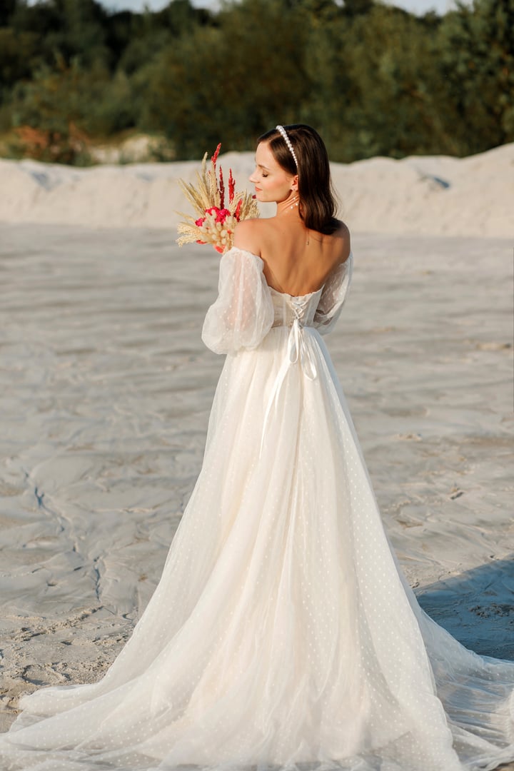 A pretty brunette girl in a white dress stands casually with a bouquet of dry flowers on the background of a white sand quarry, wedding concept