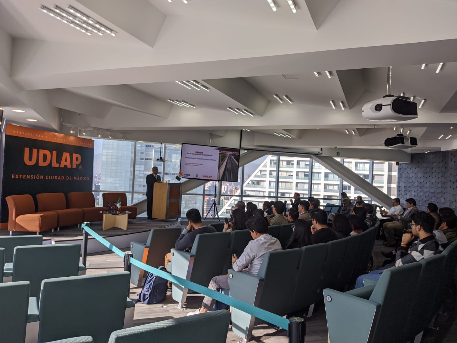 Conference room with attendees seated, facing a speaker. UDLAP banner present, indicating a university event.