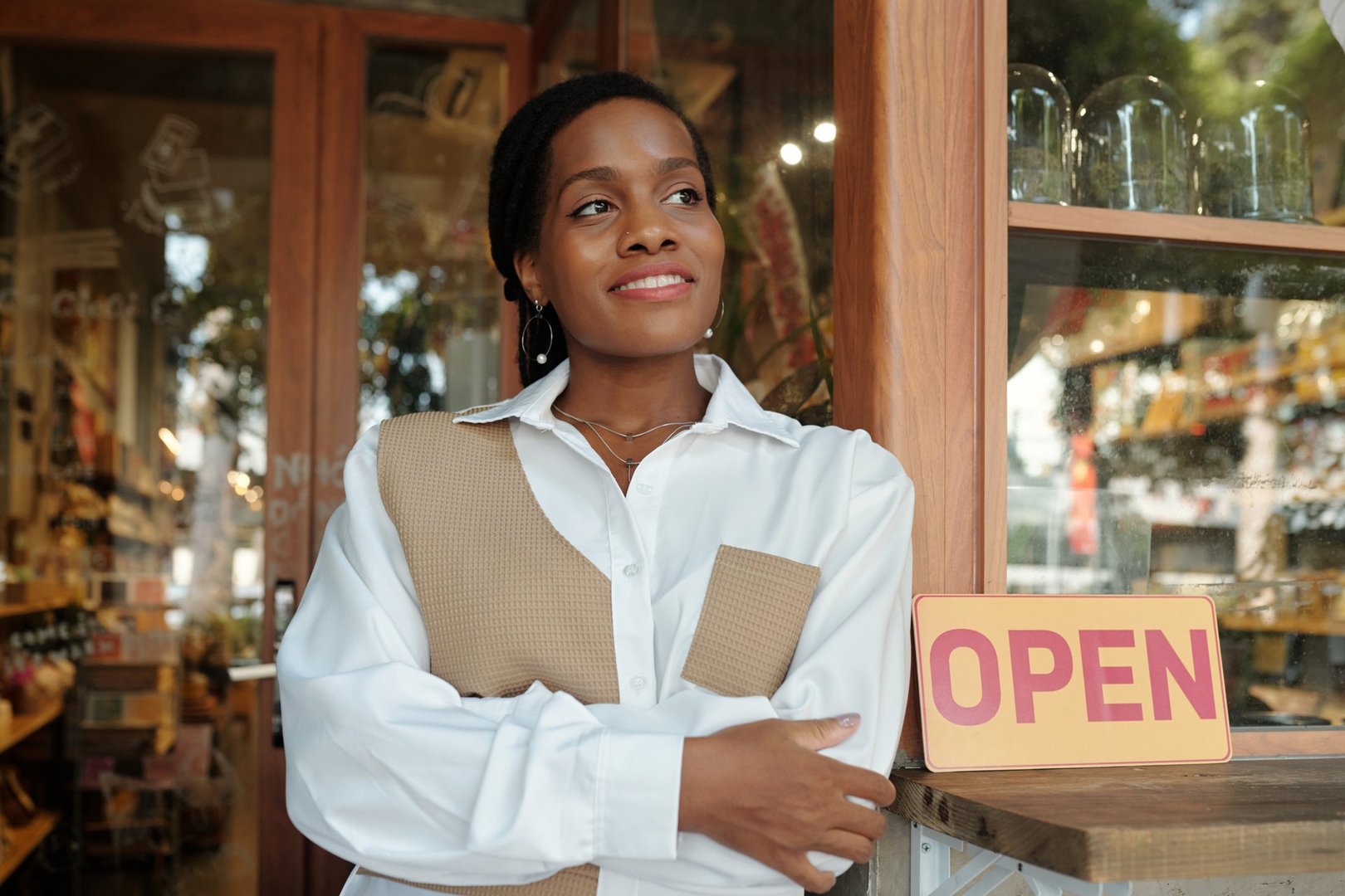 Woman standing outside her store with an open sign, smiling and folding arms wearing a beige vest over a white shirt, showing confidence and pride in her business