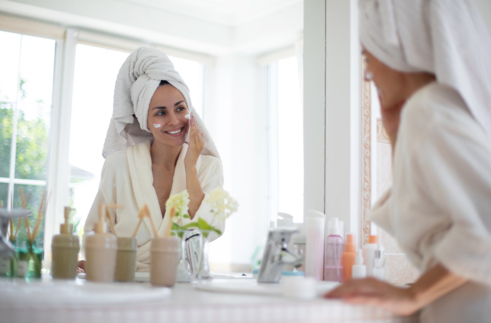 Smiling woman in white robe applies skincare cream while looking at herself in bathroom mirror. Her hair is wrapped in towel, surrounded by beauty products and plants, evoking fresh, relaxing vibe