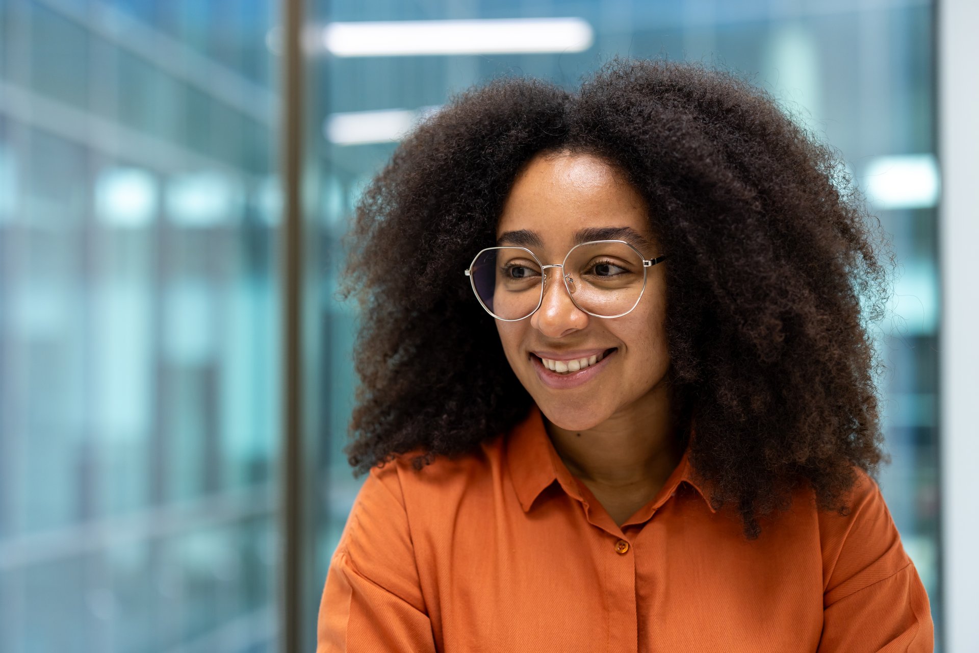 Portrait of a cheerful young woman with glasses, sitting in a modern office setting.