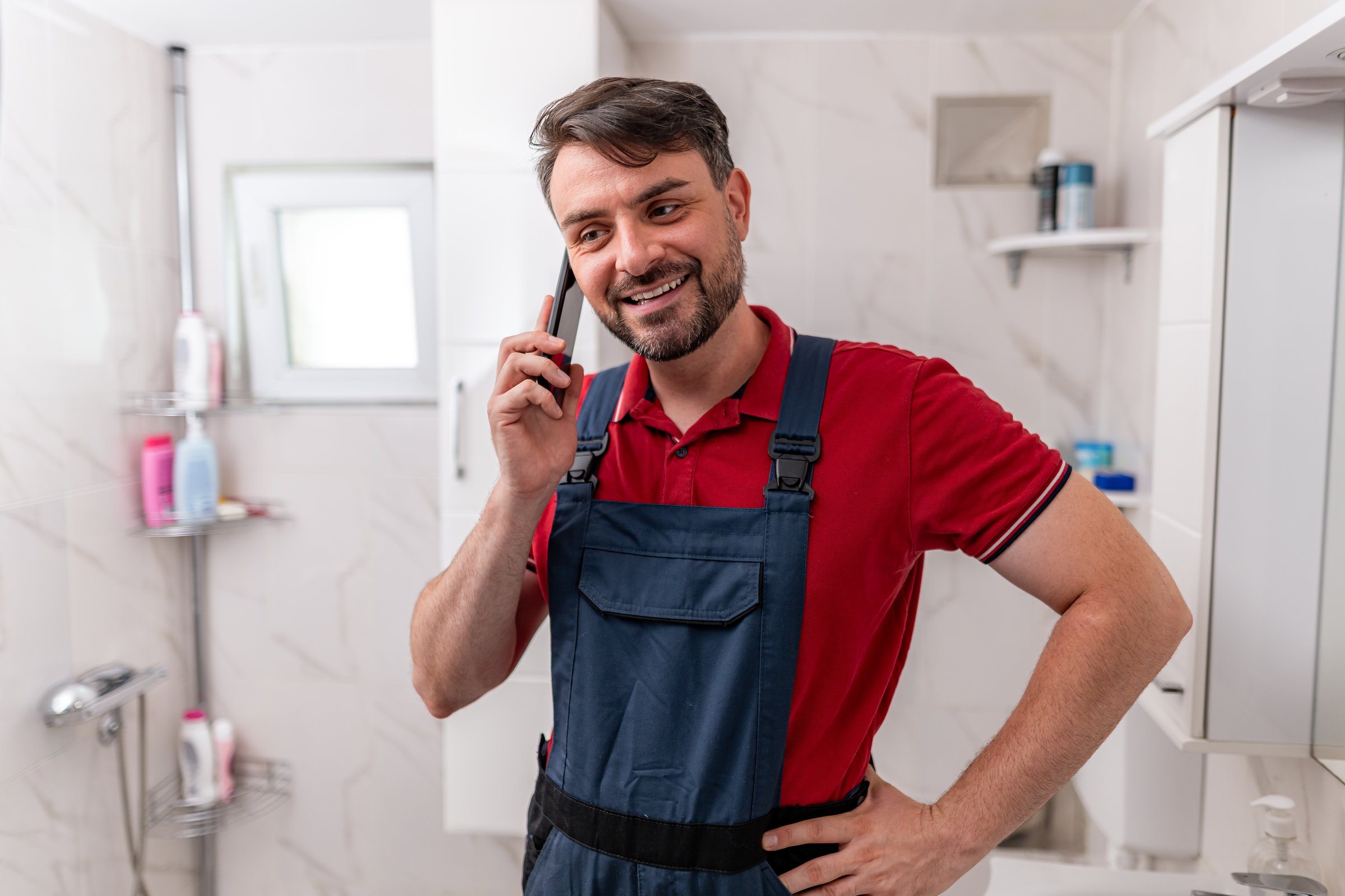 A plumber in a red shirt and blue overalls smiles while talking on the phone inside a bathroom. He seems engaged and ready for work.