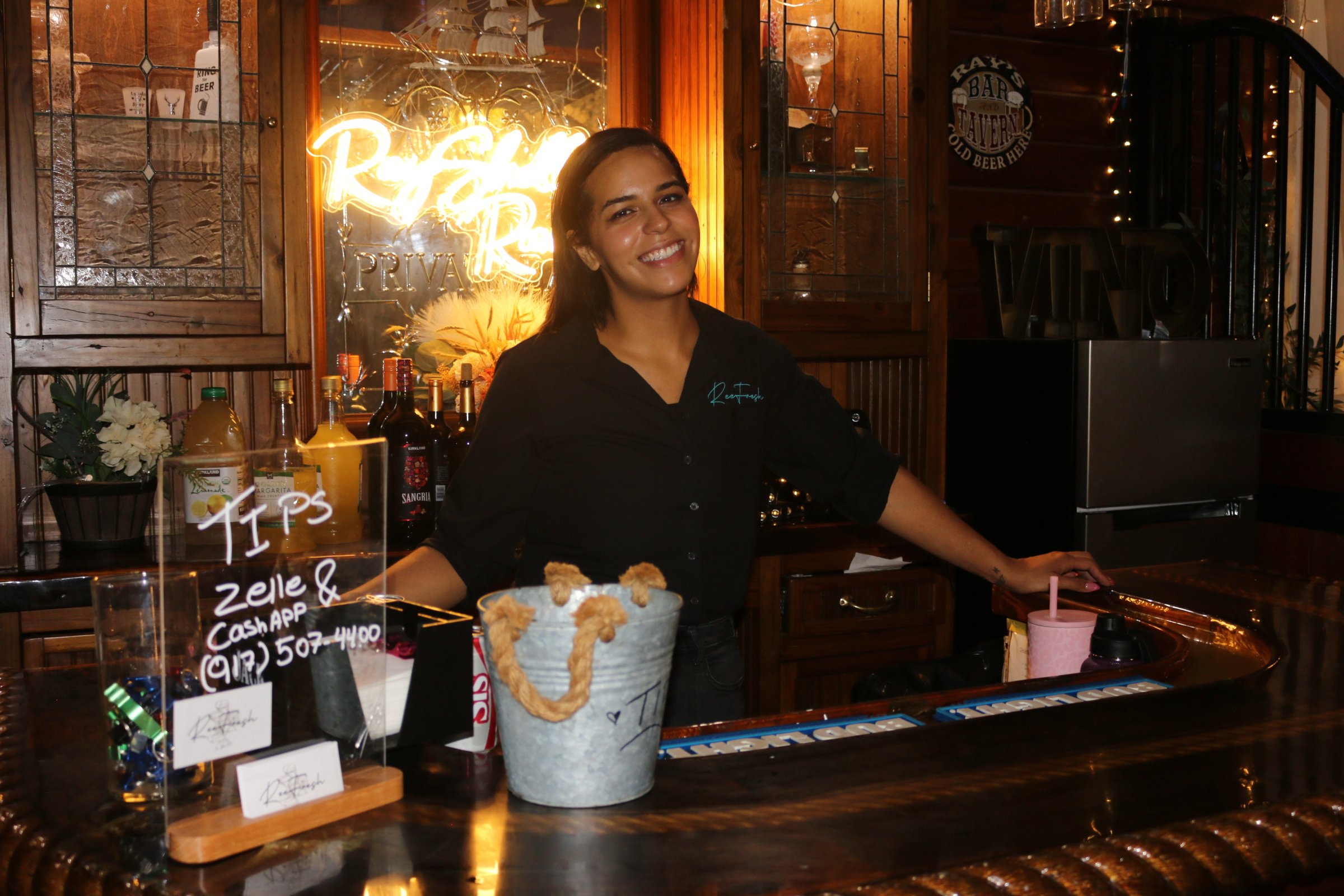 Person smiling behind a bar counter with drinks, plants, and decorative lights in the background.
