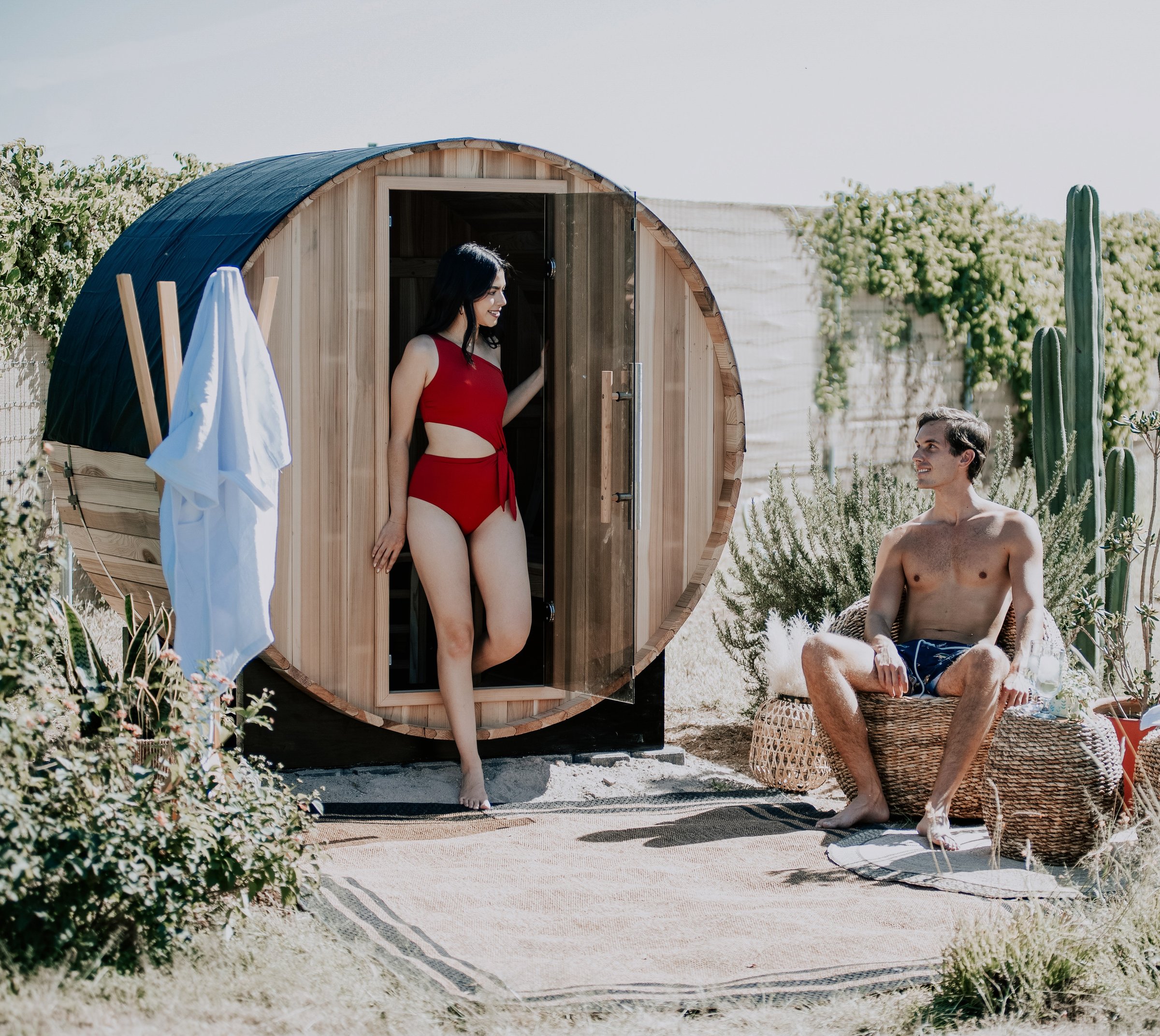 Woman in red swimsuit standing in sauna doorway, man sitting outside on wicker chair, surrounded by greenery.