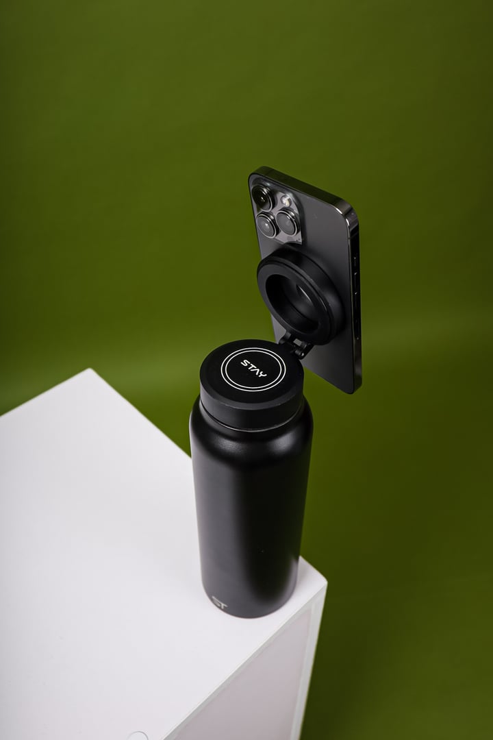 A black water bottle on a white surface with a smartphone attached to its lid against a green background.