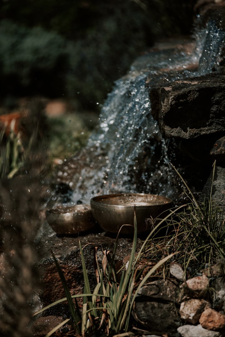 Meditative brass bowls placed beside a small cascading waterfall in a natural outdoor setting.