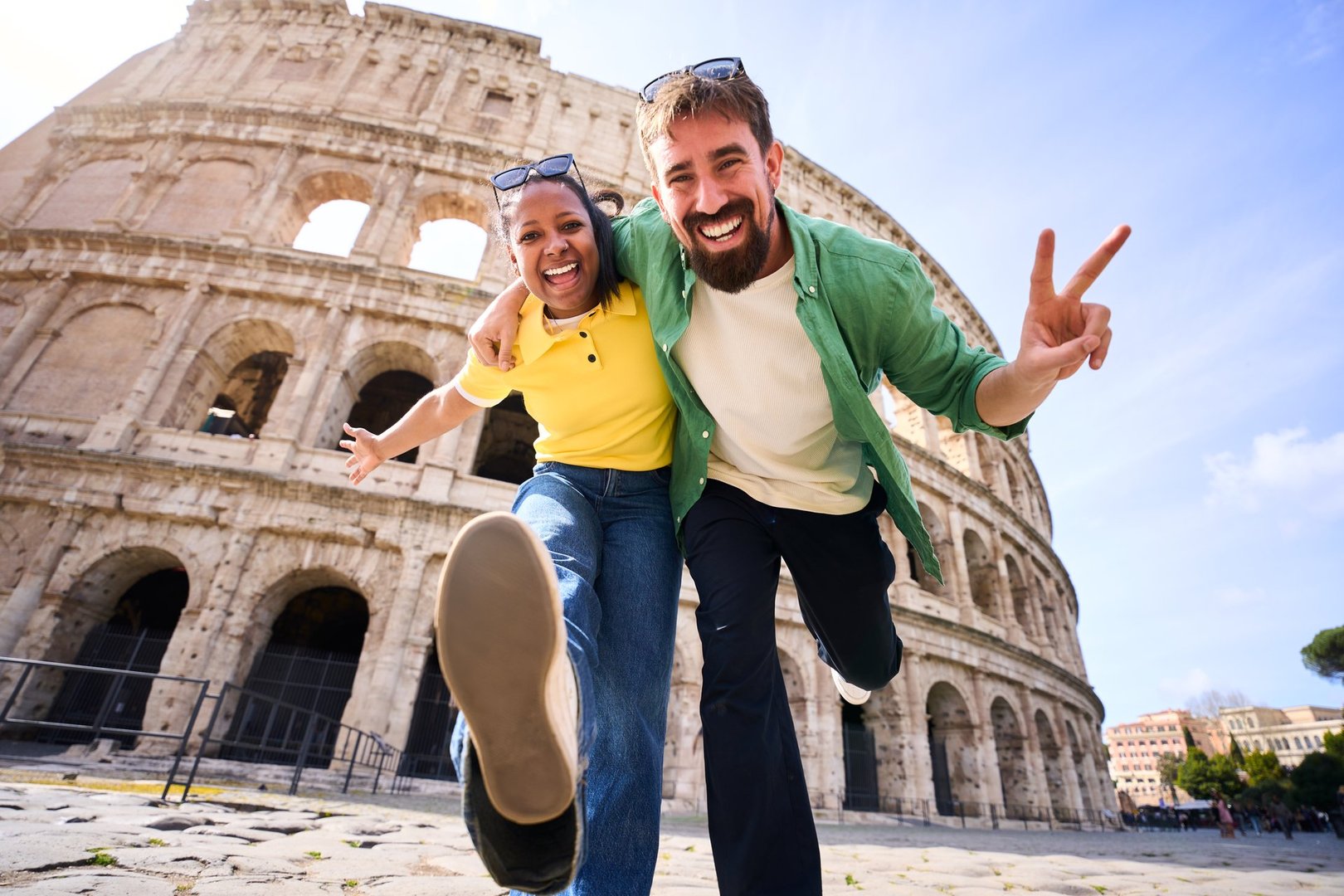 Happy multi-ethnic couple of tourists visiting the colosseum in rome, italy, joyfully making victory signs while smiling and enjoying their memorable adventure under the sunny sky