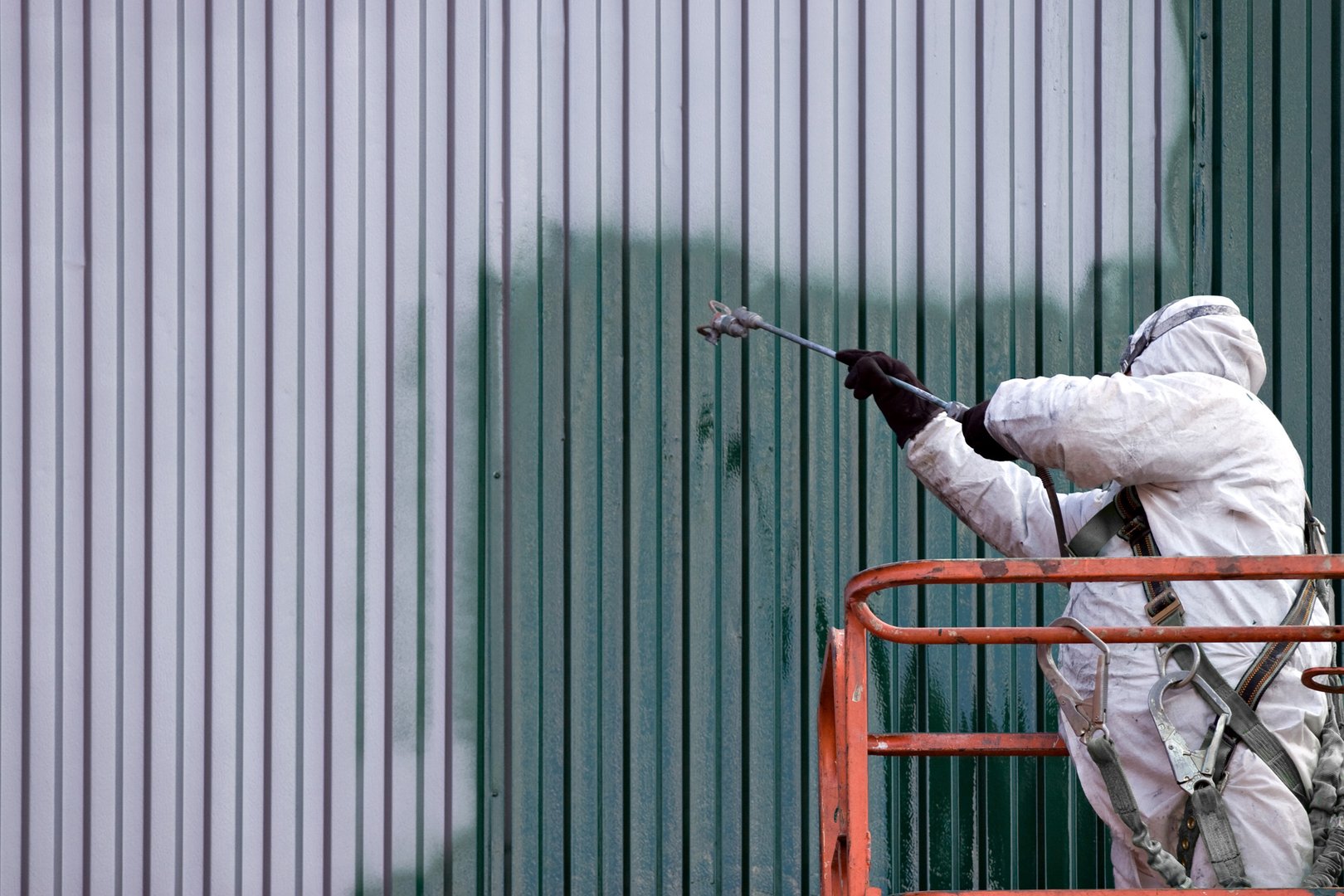 Person in protective gear spray-painting a corrugated metal wall green from an elevated platform.