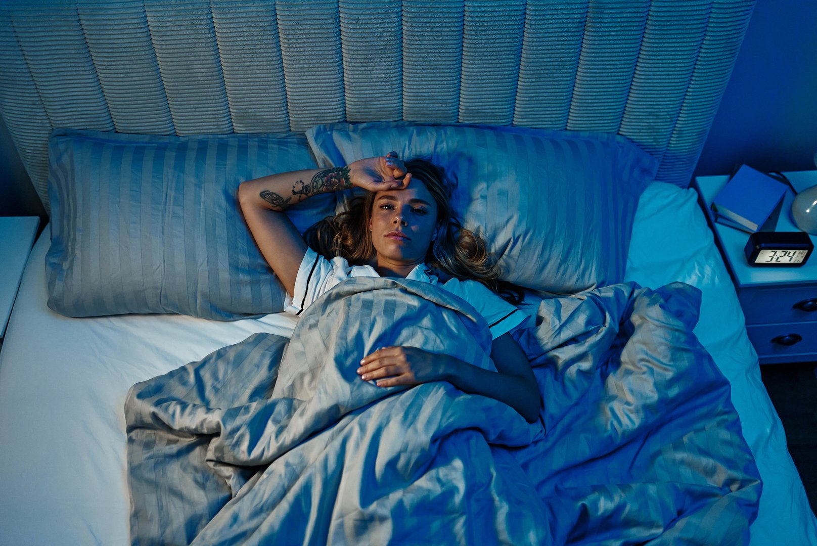 A woman lies awake in bed, showing signs of insomnia and anxiety despite the calming, modern bedroom around her. She struggles to relax amid cozy comforts, affecting her health and wellness