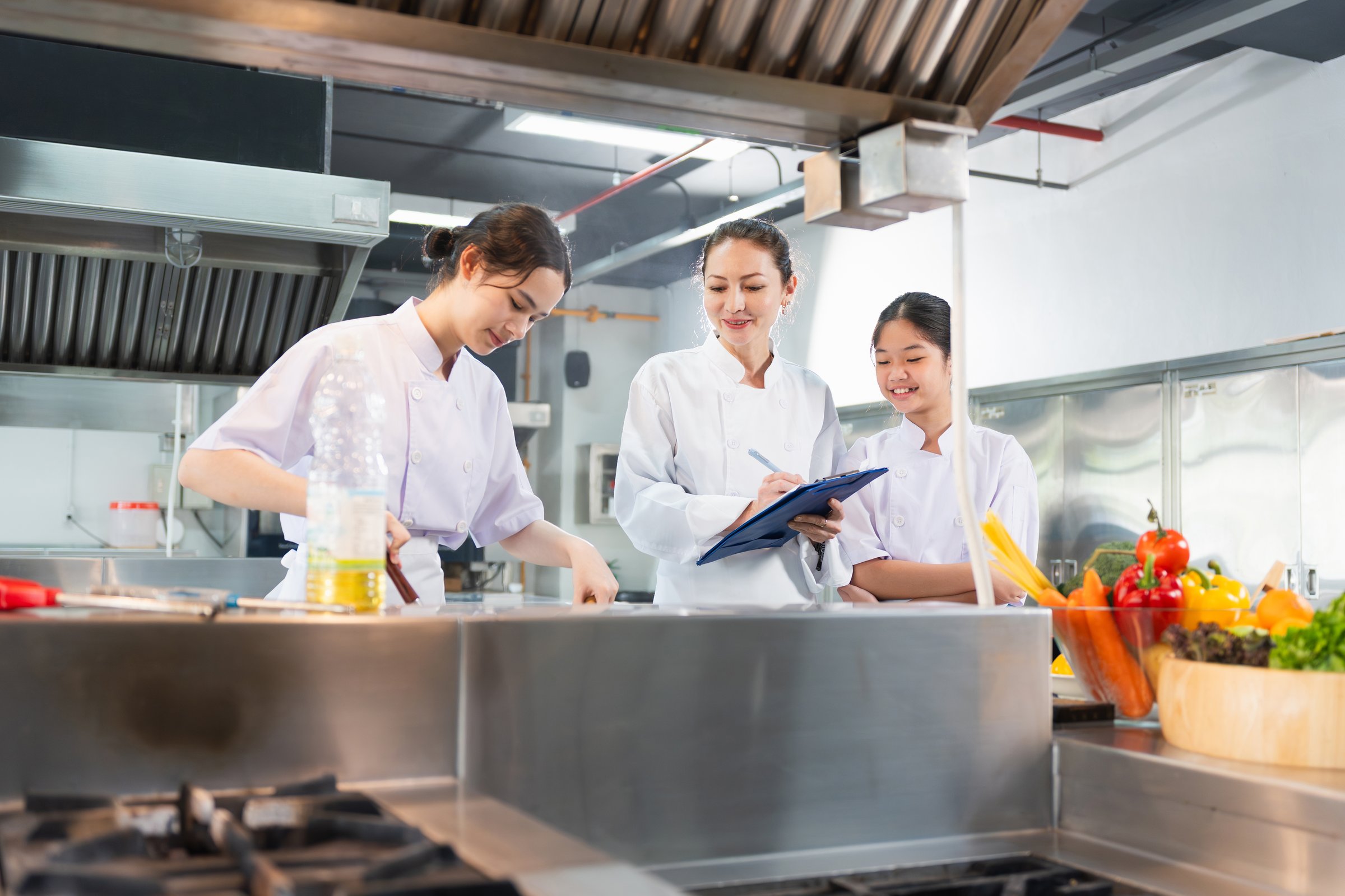 Culinary Team Collaborating and Learning in Restaurant Kitchen, Female Chefs Preparing Food and Taking Notes in a Modern Kitchen, Professional Chefs Working Together in a Busy Commercial Kitchen