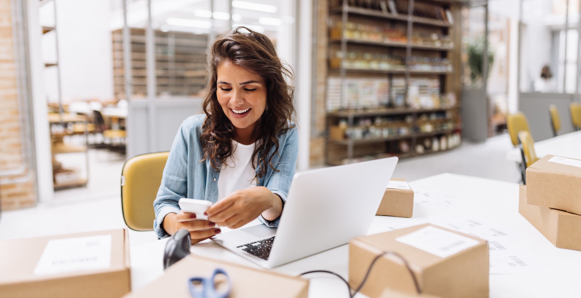 Smiling online store owner reading a text message on her smartphone. Happy businesswoman making plans for product shipping in her warehouse. Female entrepreneur running an e-commerce small business.