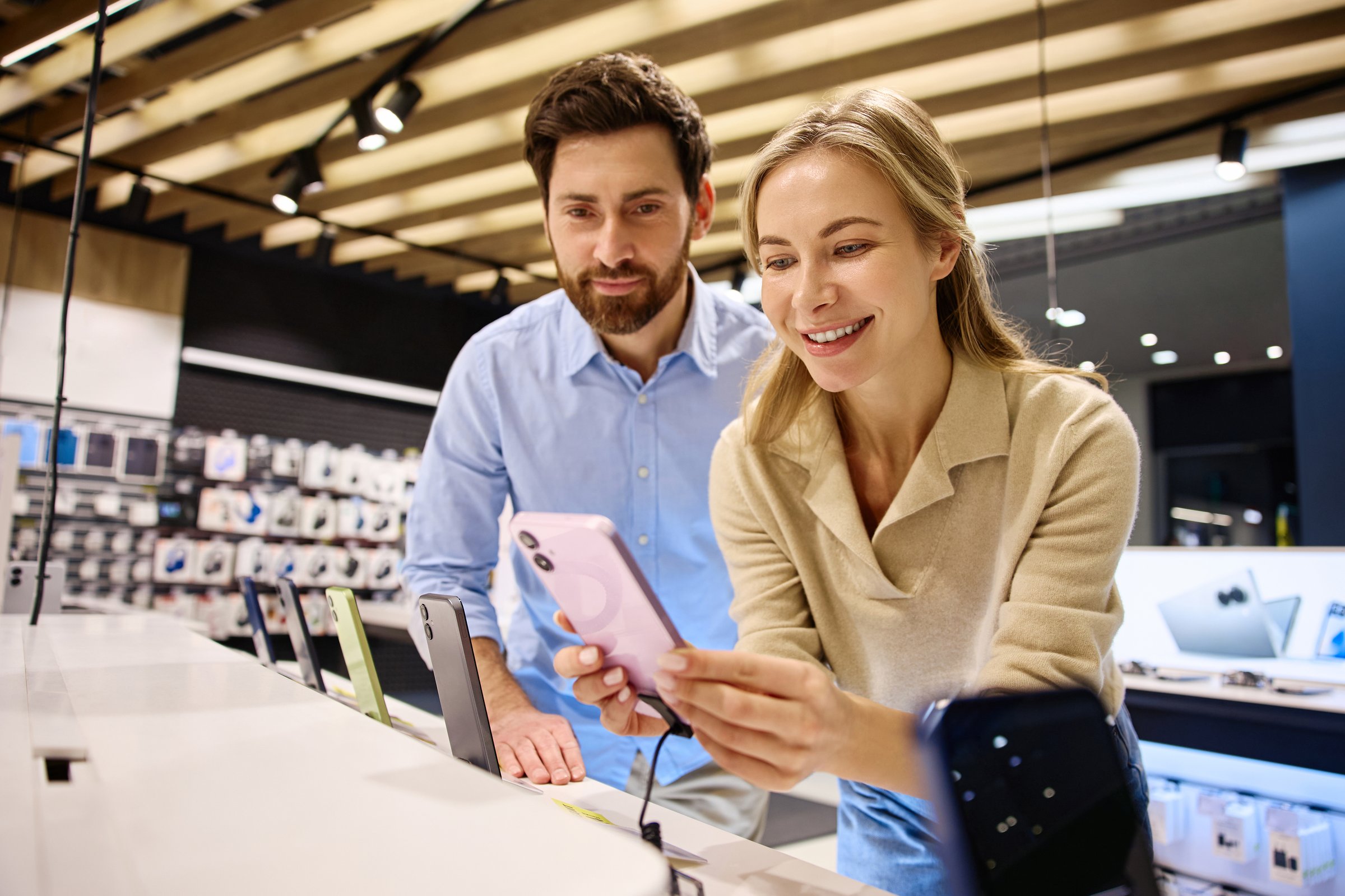 In a busy electronics store, a man and woman browse the latest smartphones, sharing their excitement for modern tech and trends that captivate consumers, making them feel youthful and engaged