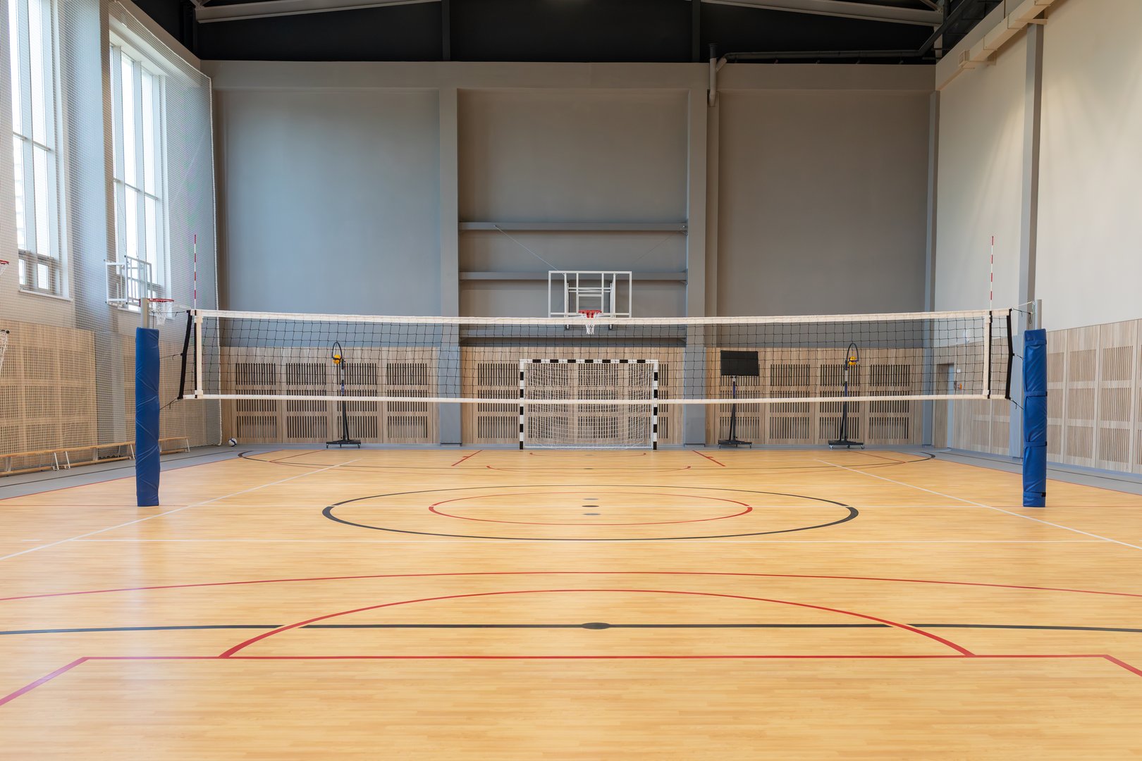 Indoor sports hall with volleyball net and basketball hoop on wooden floor. Multi-purpose court for school or community recreation. Real photo.