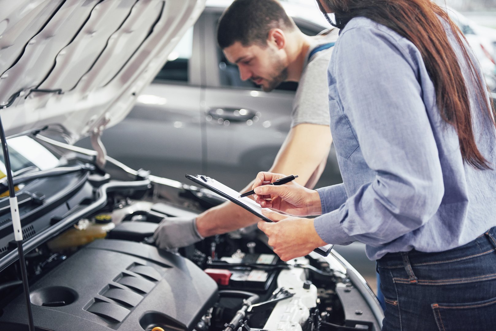 Man mechanic and woman customer looking at car hood discussing repairs