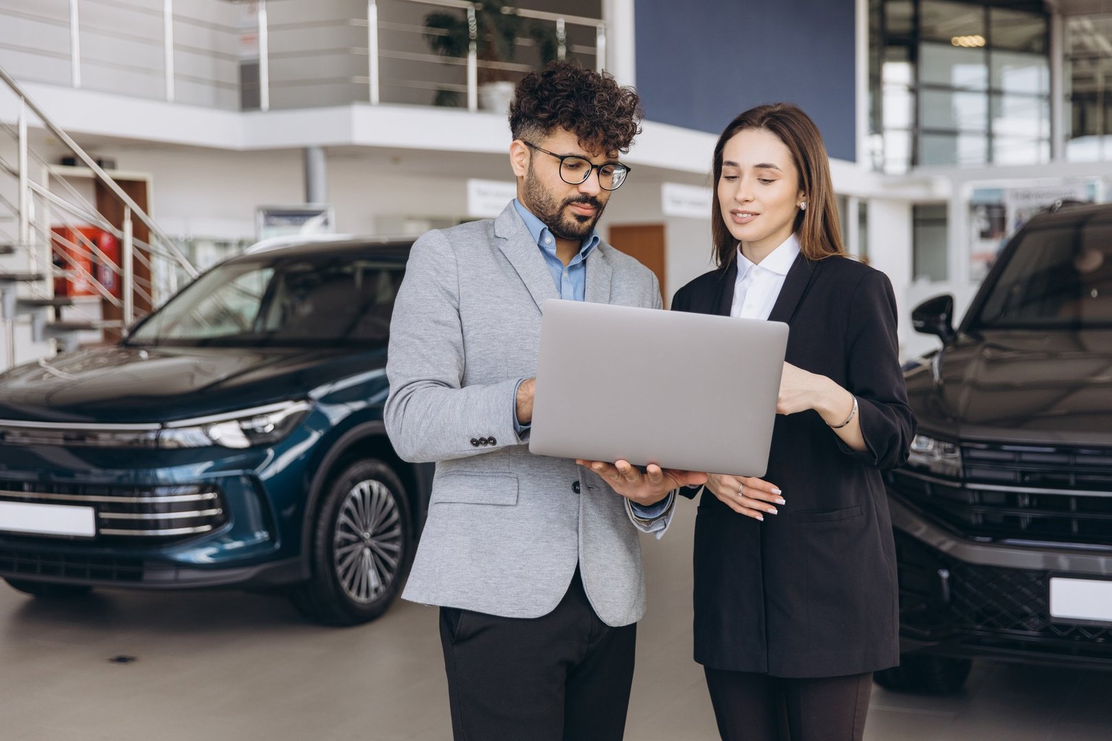 Salesman presenting vehicles on a laptop to a customer in a vibrant car dealership showroom, facilitating the buying process