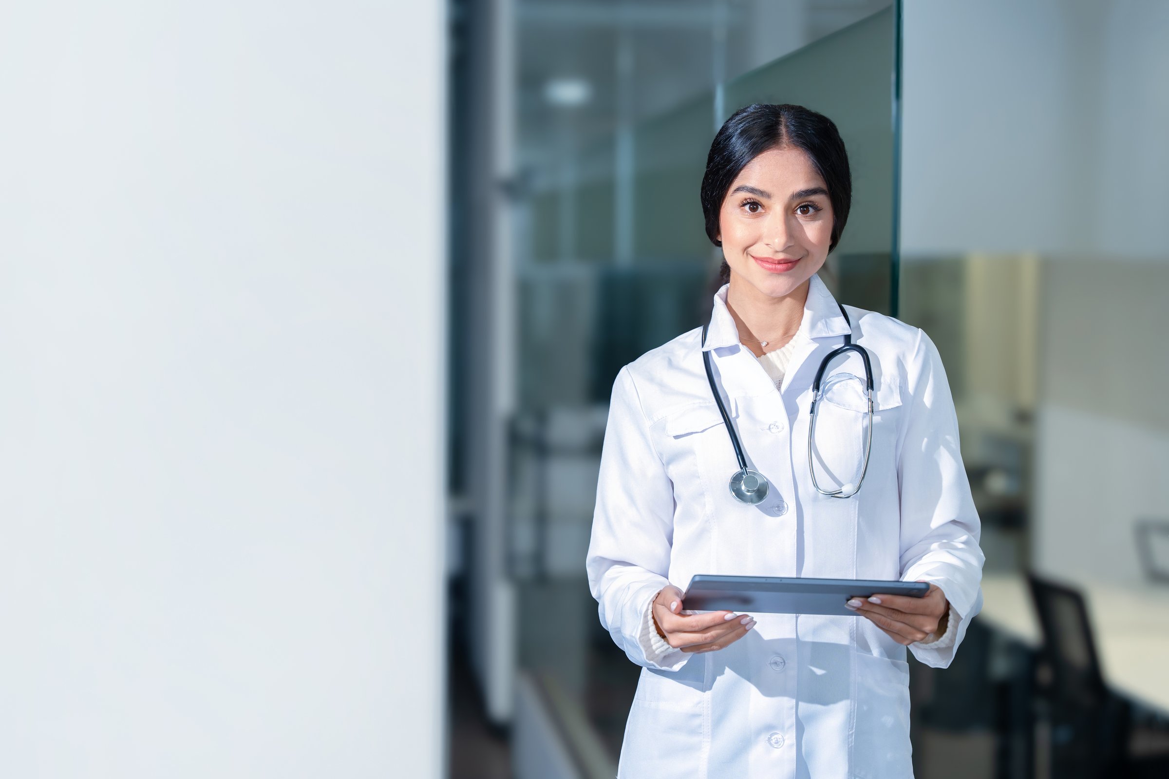 Portrait of beautiful Indian female doctor wearing white lab coat, using tablet computer while standing in front of hospital. She looks professional and confident. Background with copy space.