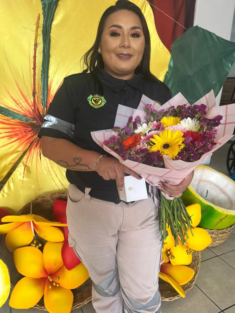 Woman in uniform holding a colorful bouquet, standing in front of floral backdrop, smiling at the camera.