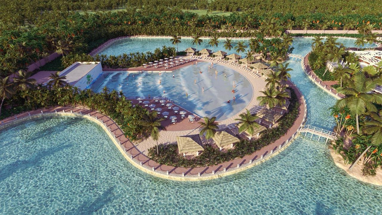 Aerial view of a tropical resort pool with palm trees, sun loungers, and people swimming, surrounded by lush greenery.