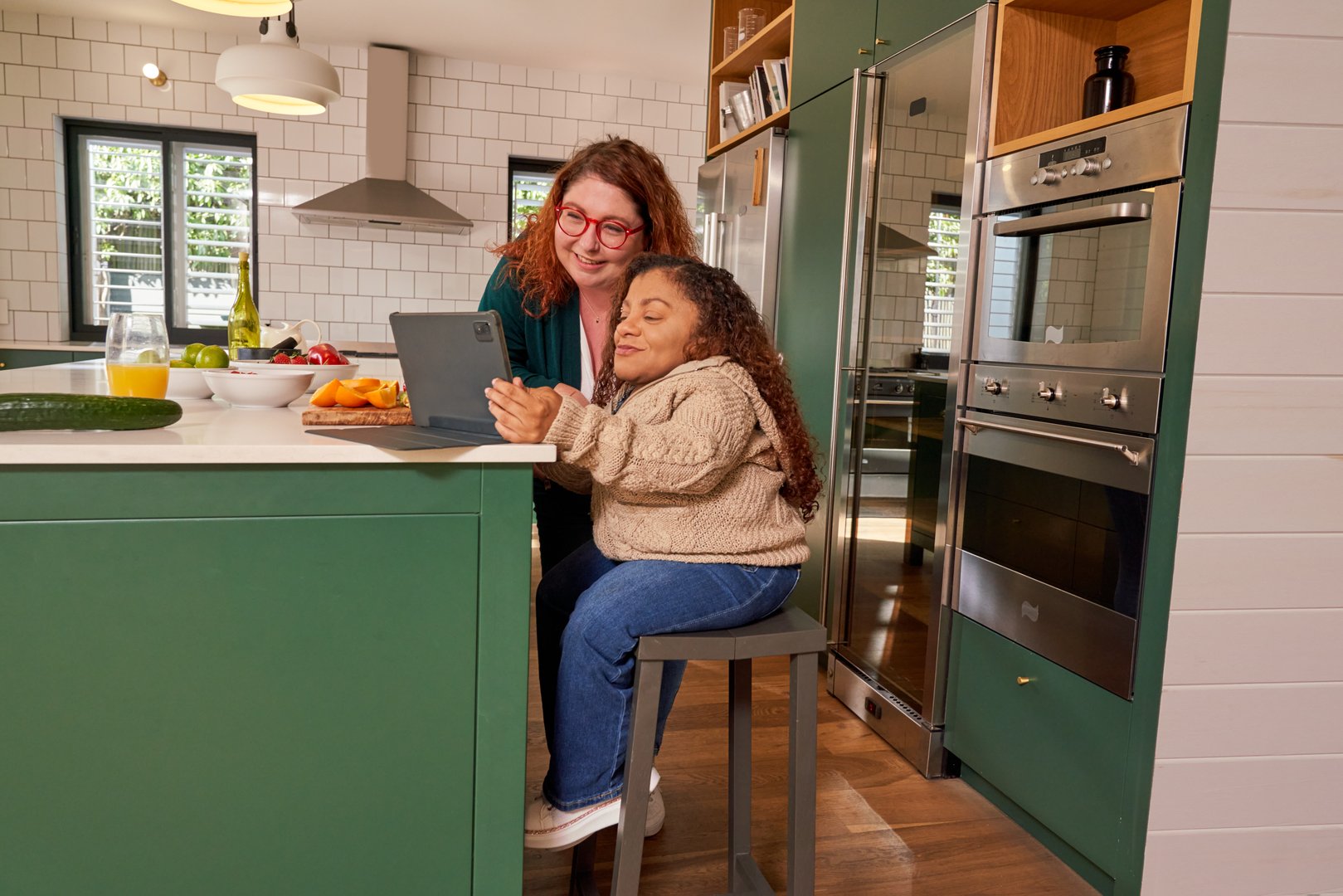 Woman assisting friend with osteogenesis imperfecta using tablet in kitchen