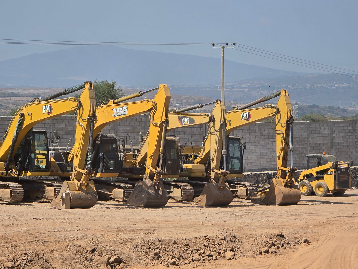 Four yellow excavators lined up on a construction site with a mountain in the background.