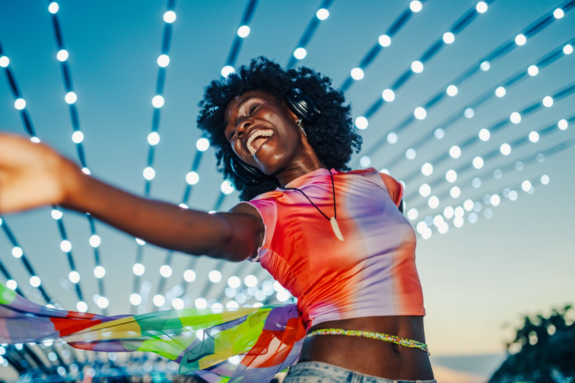 Carefree young woman enjoying music and dancing with a colorful scarf under string lights at an outdoor summer music festival