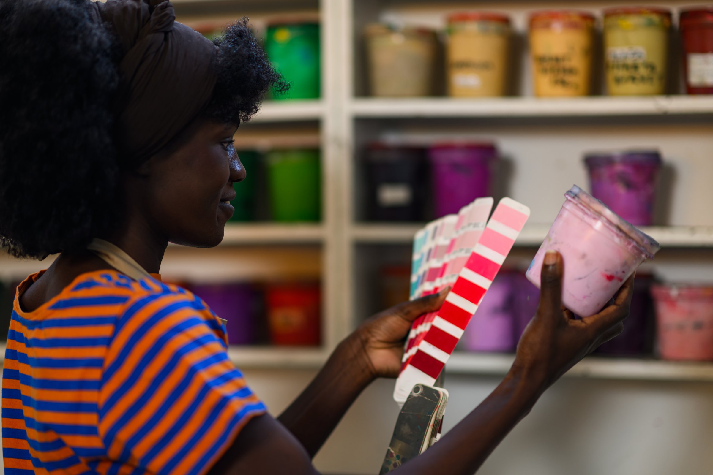 Profile view of multicultural female graphic worker standing at printing workshop with ink bucket and color swatch in hands and choosing right color. Smiling diverse graphic technician with ink bucket