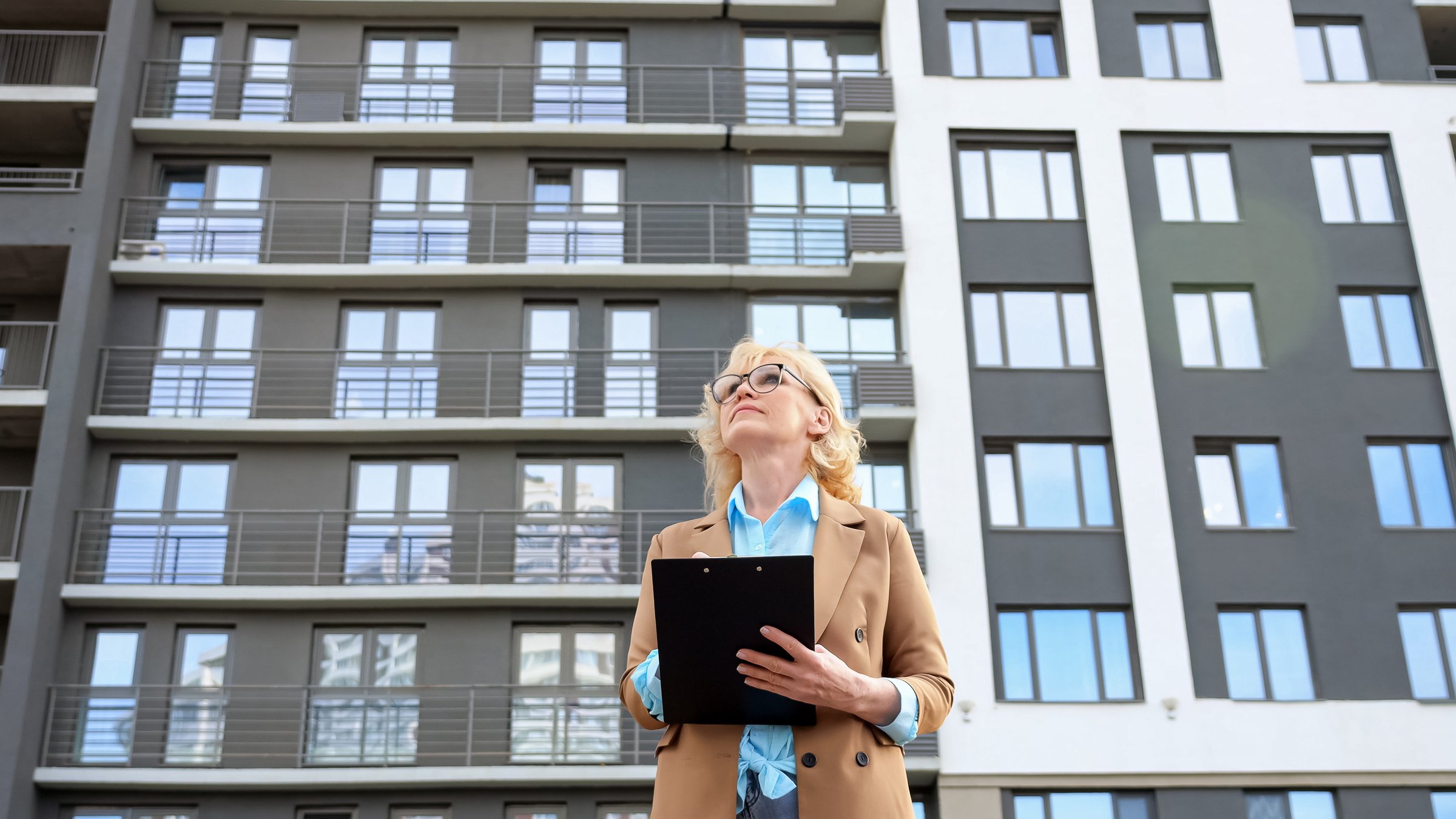 Senior woman entrepreneur looks around making notes on clipboard on apartment building background. Concept of working as real estate agent low angle shot