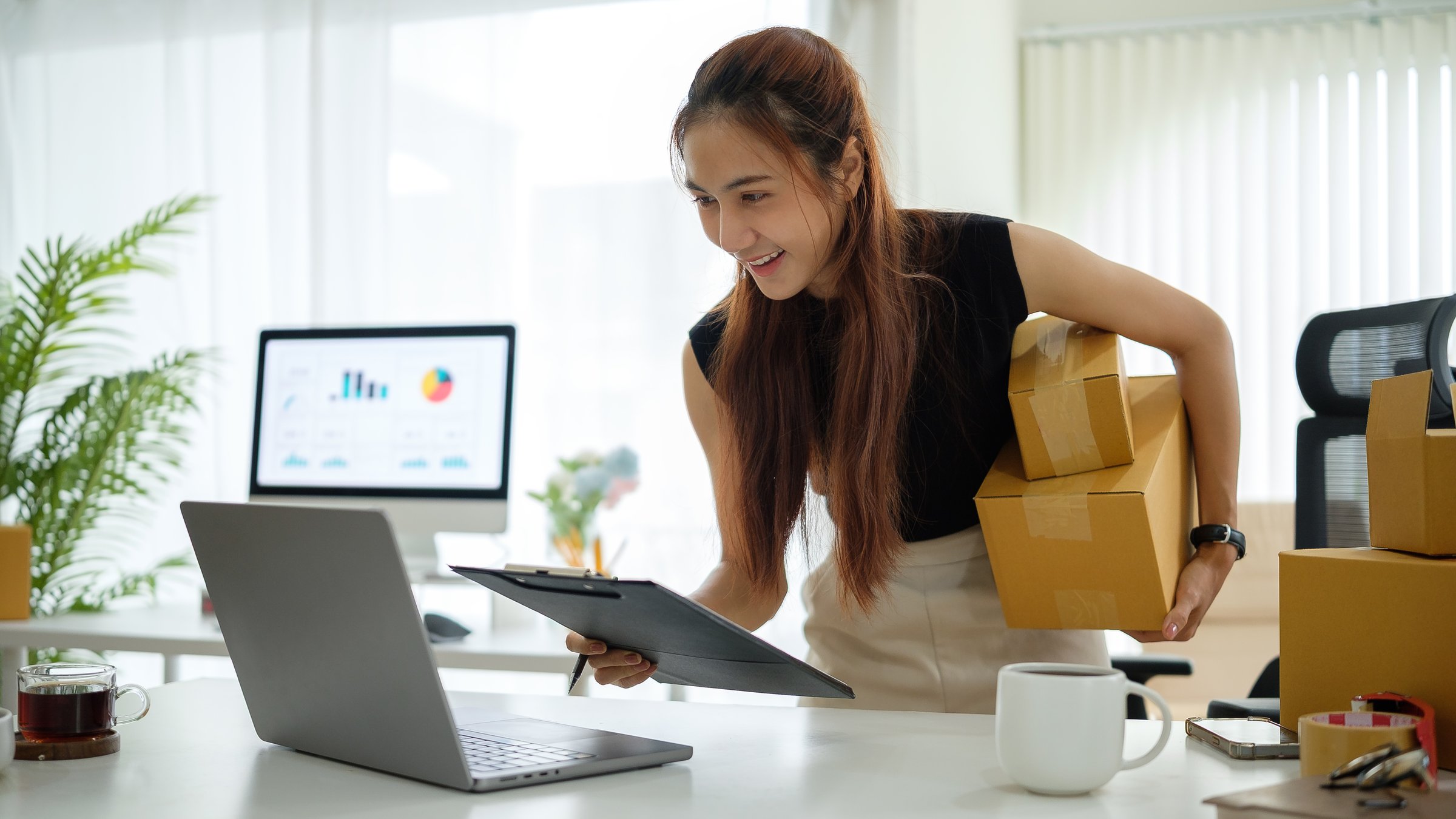 Confident young woman holding shipping boxes and checking order details on a laptop at her modern home office.