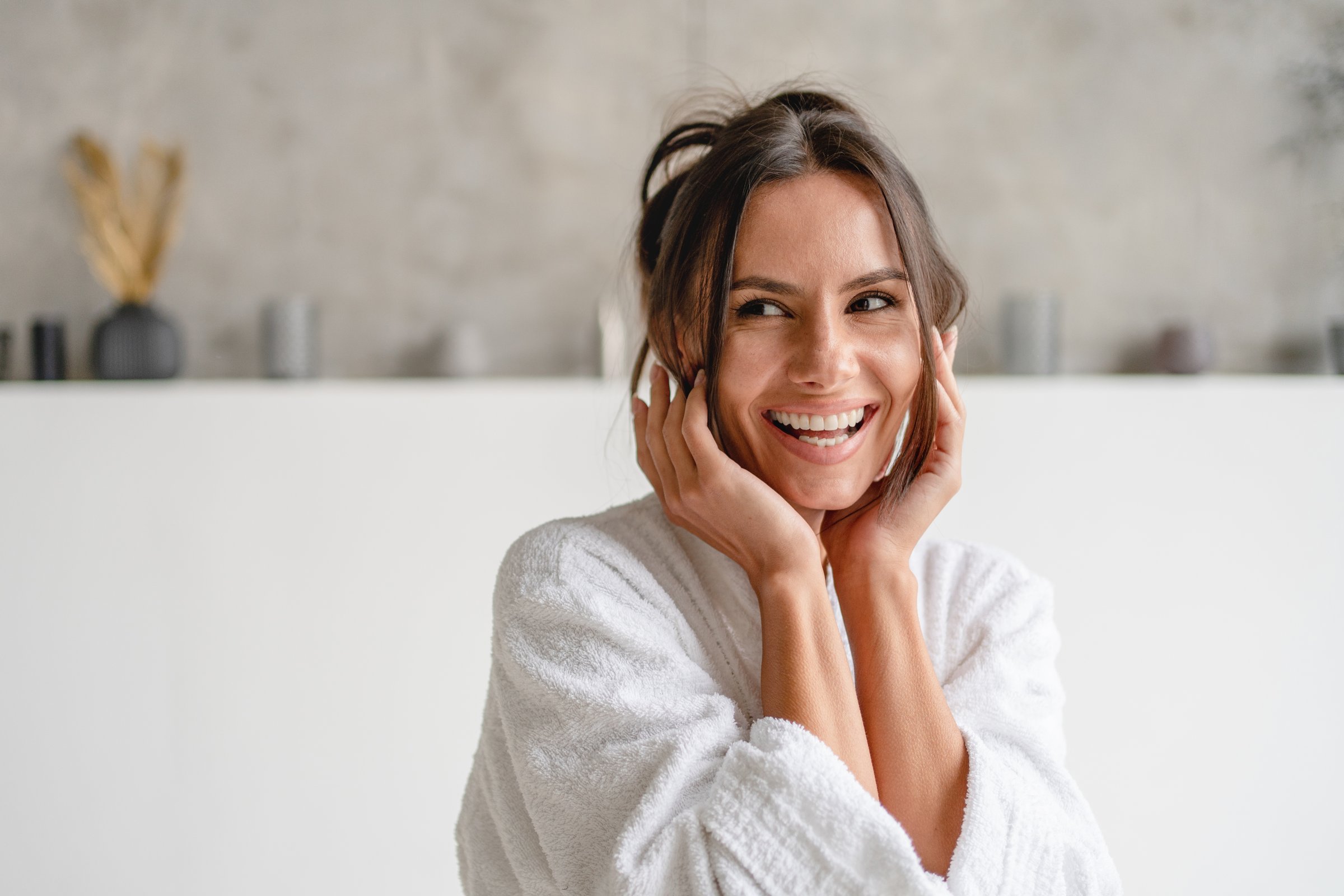 Beautiful young Caucasian brunette lady in bathrobe enjoying sunny morning in luxury bathroom touching her face fresh glowy skin, looking aside away on copy space. Wellness beauty routine concept