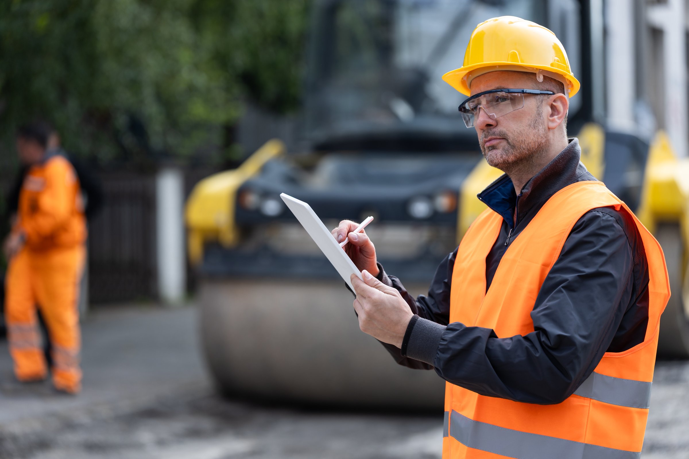 A construction site supervisor reviews plans on a tablet, dressed in safety gear with a bright orange vest and helmet. Workers in the background are actively engaged in their tasks.