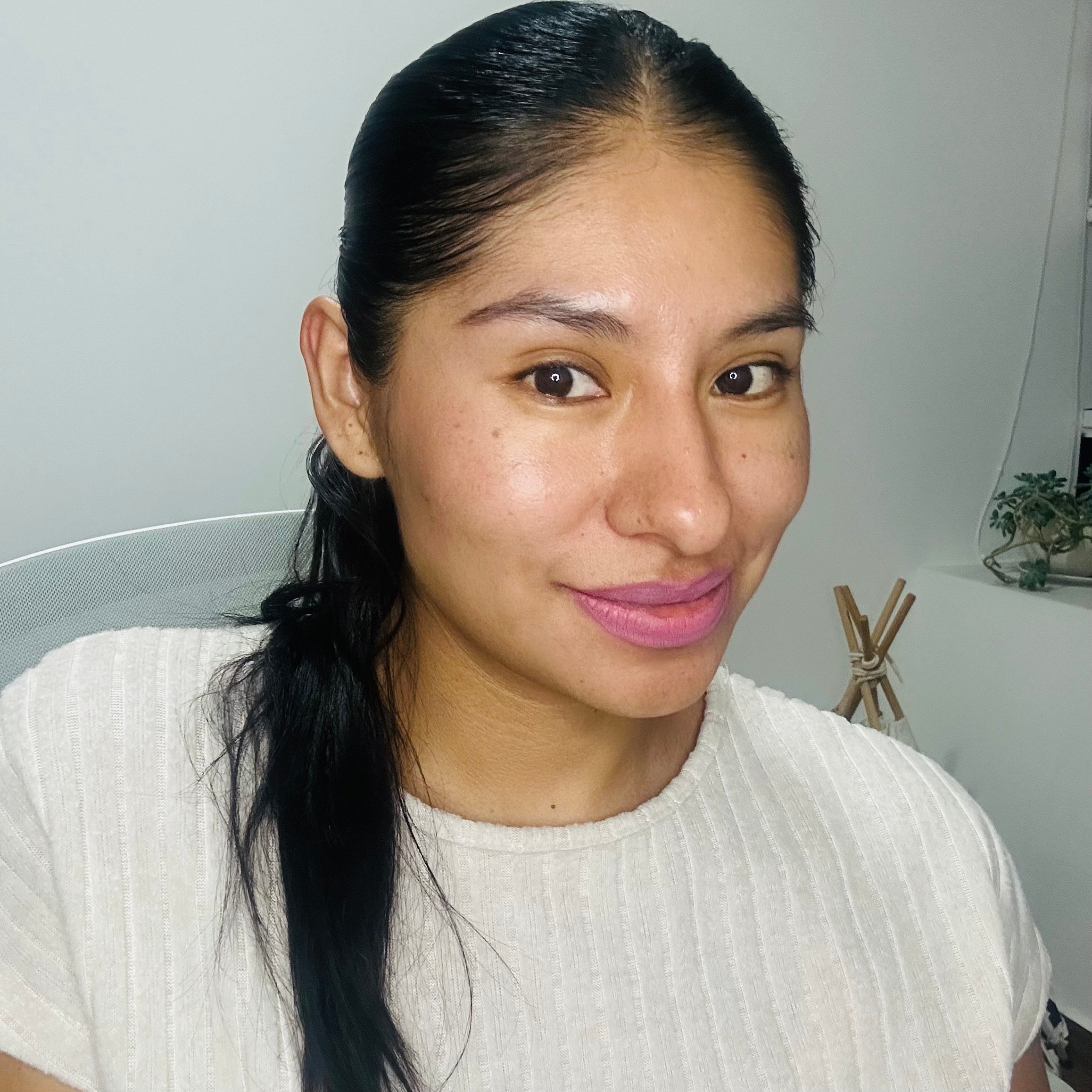 Woman with long dark hair smiles, wearing a light textured blouse, sitting indoors with a small plant in the background.