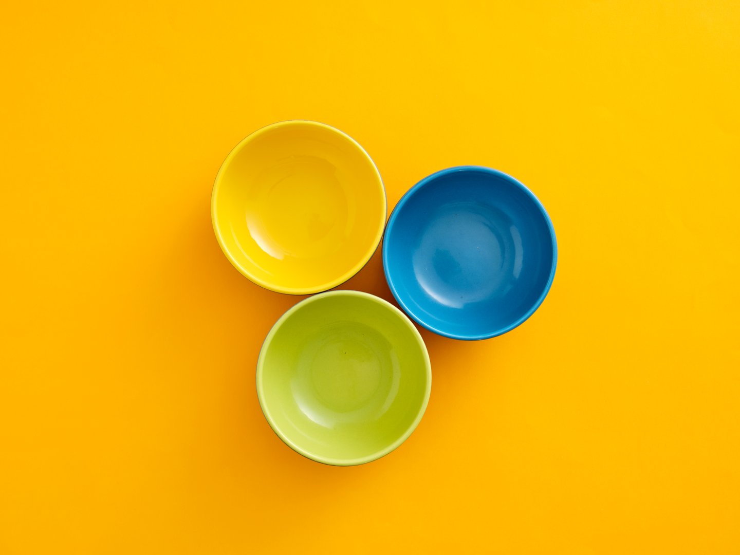 Three vibrant primary colored ceramic bowls, yellow, blue, and green, arranged on a bright yellow background, top view