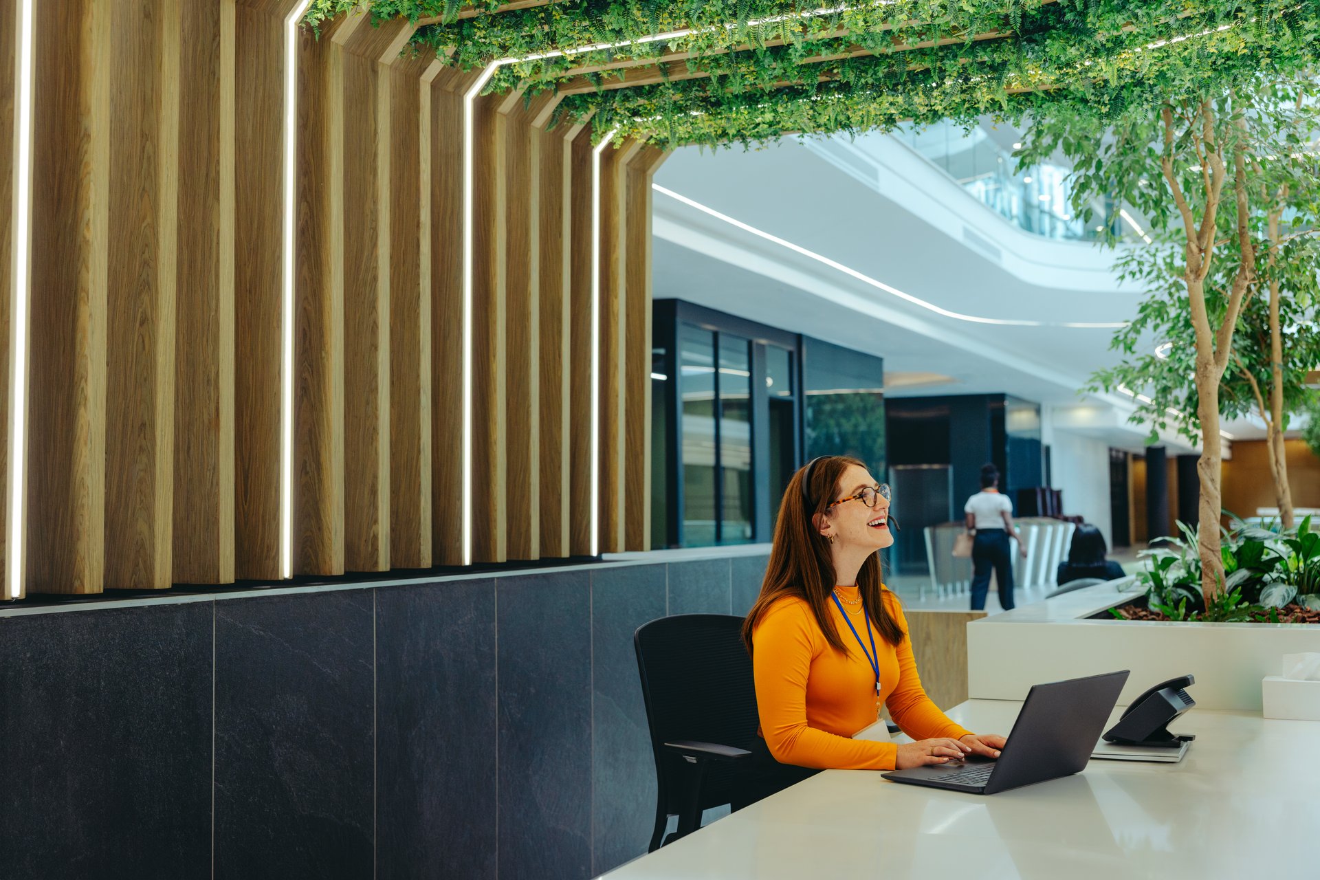 A friendly receptionist with a bright smile sits at a desk using a laptop in a modern office lobby. The setting features natural elements and contemporary design, creating a welcoming atmosphere.