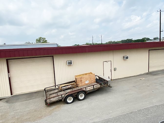 A flatbed trailer carrying a wooden crate parked in front of a beige building with closed garage doors.