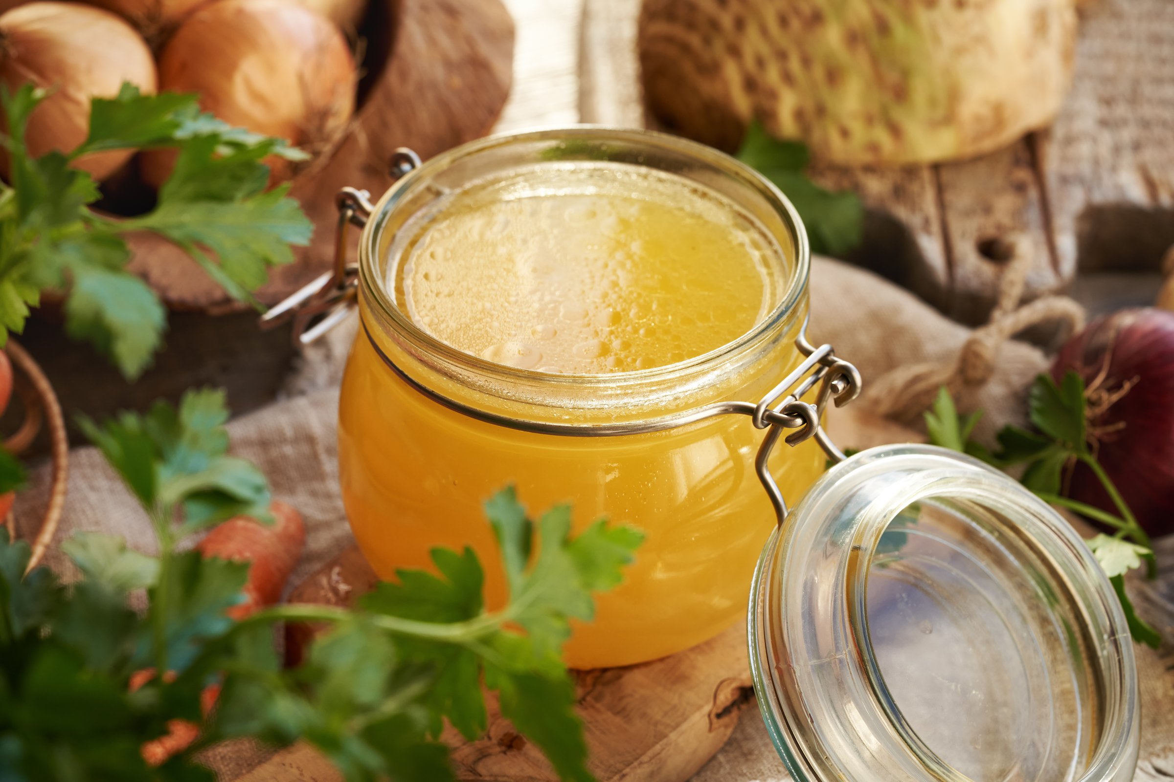 Chicken soup or bone broth in a glass jar with fresh parsley, onions and celery root in the background