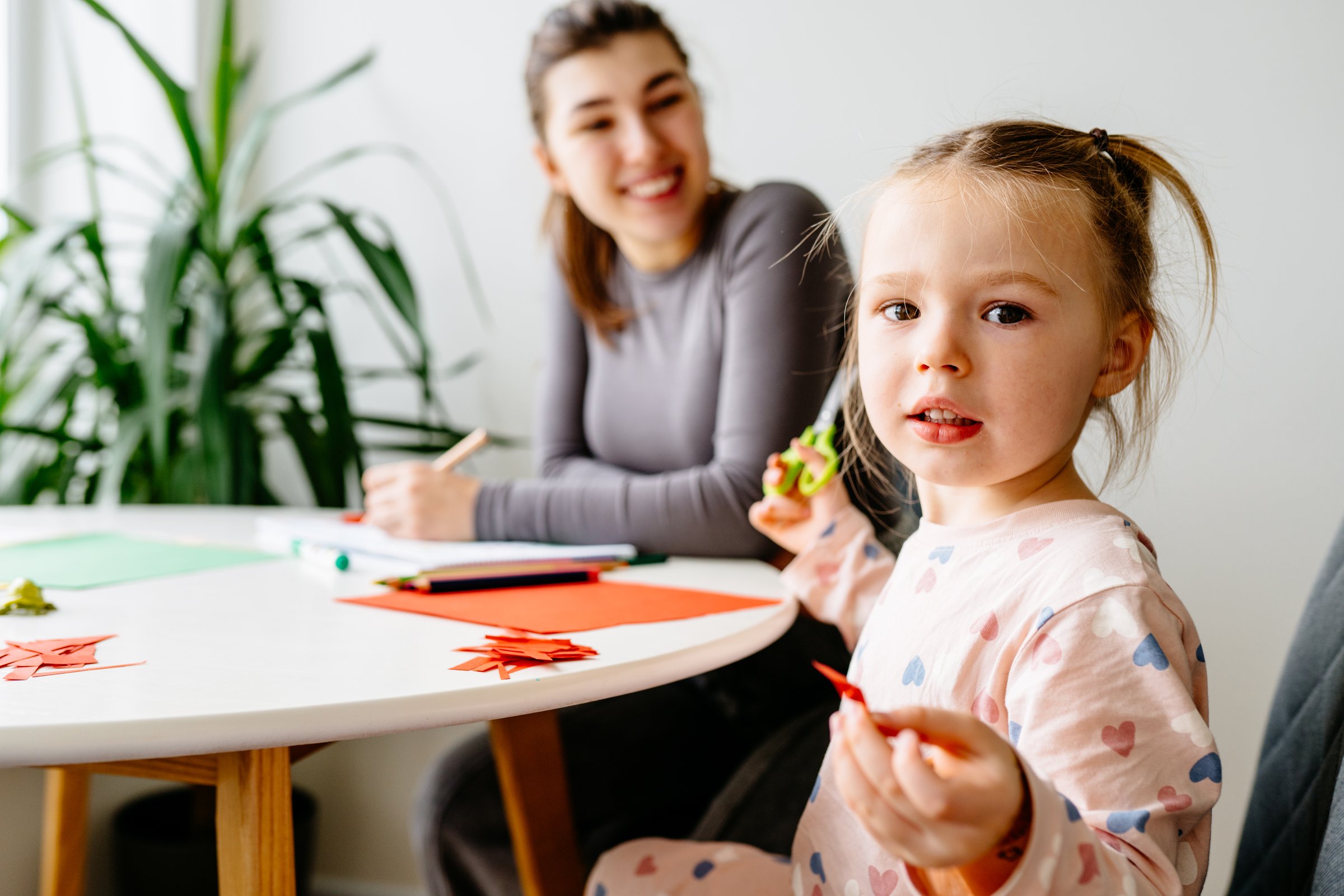 Cute girl, 3 years old, and mother do arts and crafts together, cutting shapes from colored paper while sitting in kitchen at home. Motherhood, parenting, and educational activities concept.