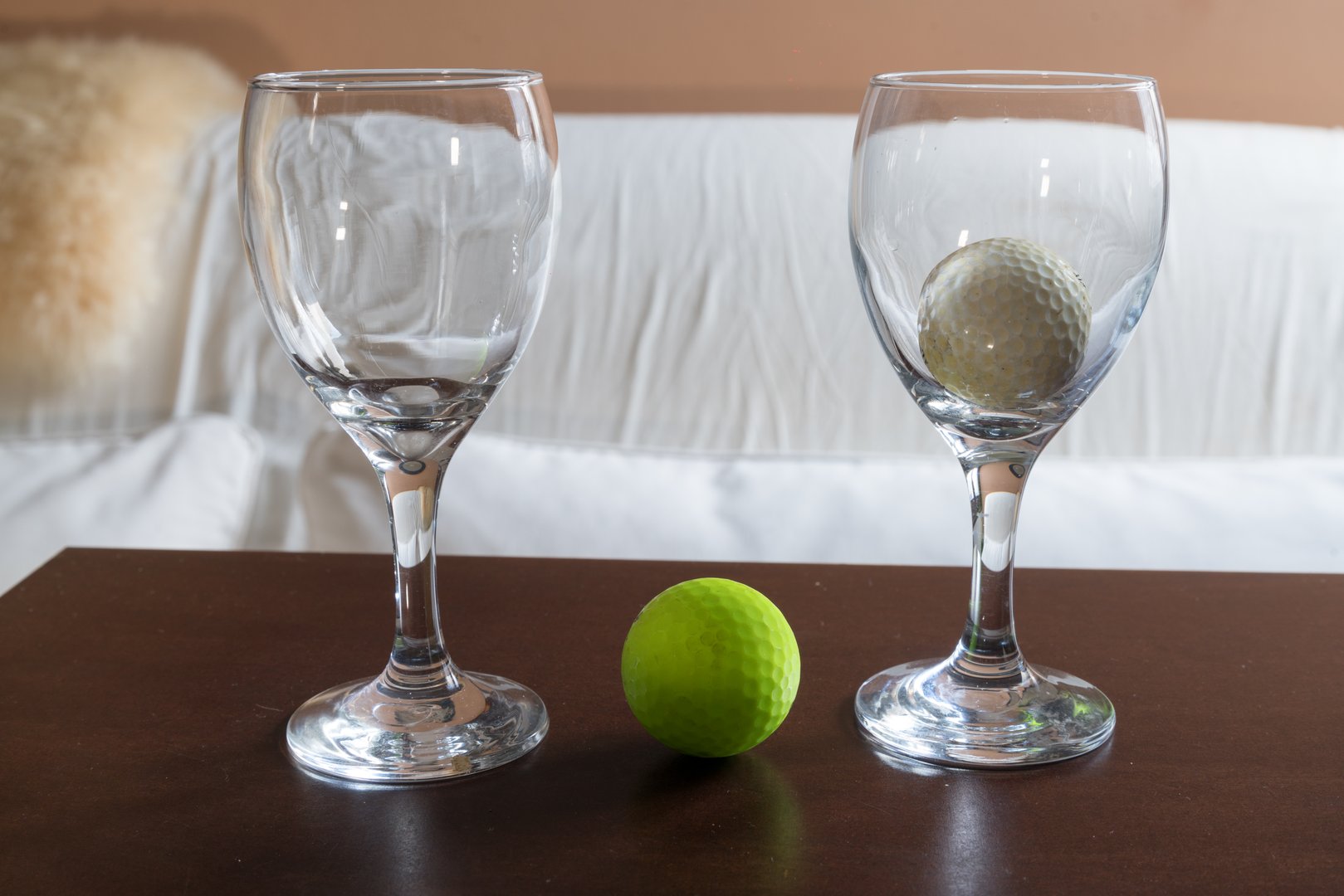 Two elegant wine glasses frame a vibrant green golf ball on a wooden table, with another golf ball placed inside one of the glasses.