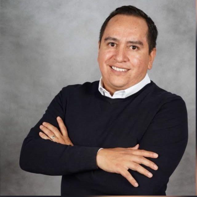 Person smiling, wearing a dark sweater and white shirt, arms crossed, in front of a gray studio background.