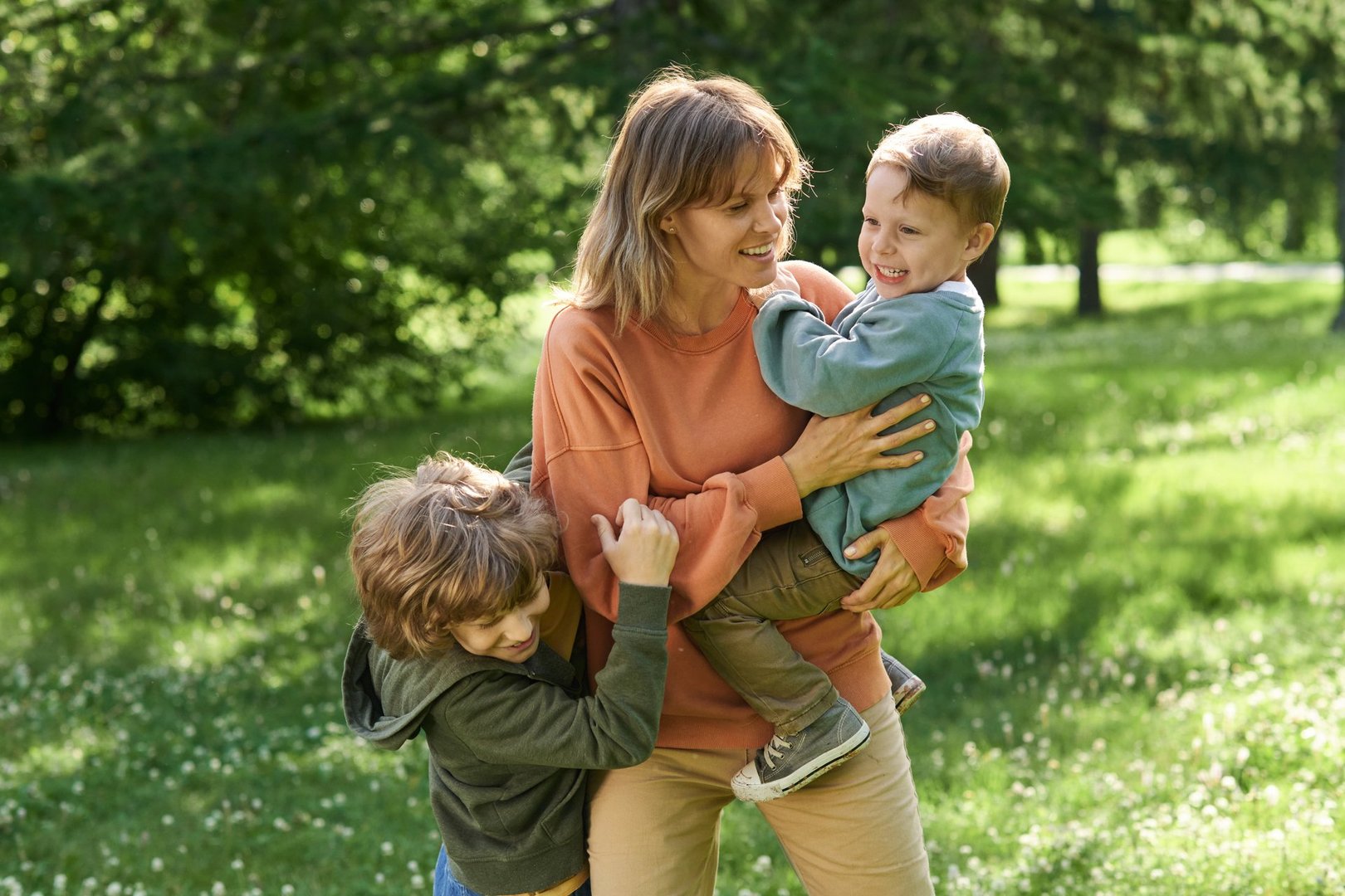 Candid scene of happy mother playing with sons outdoors in green sunlight meadow and holding child