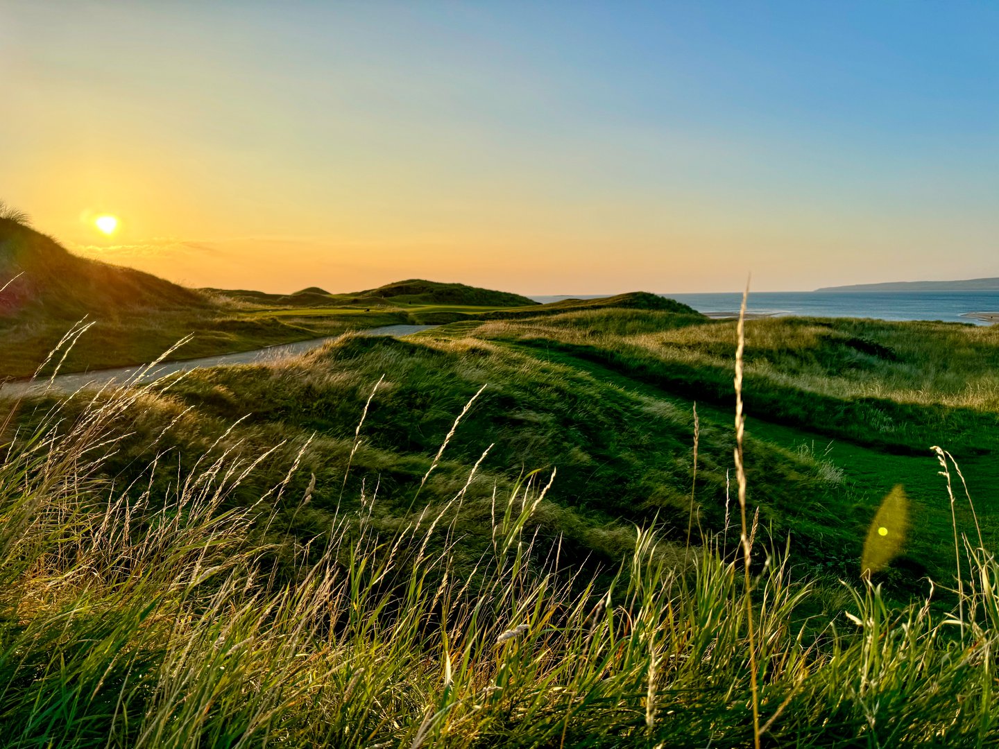 A breathtaking sunset casts vibrant colors over a sprawling grassy field, with a winding road meandering through the tranquil landscape