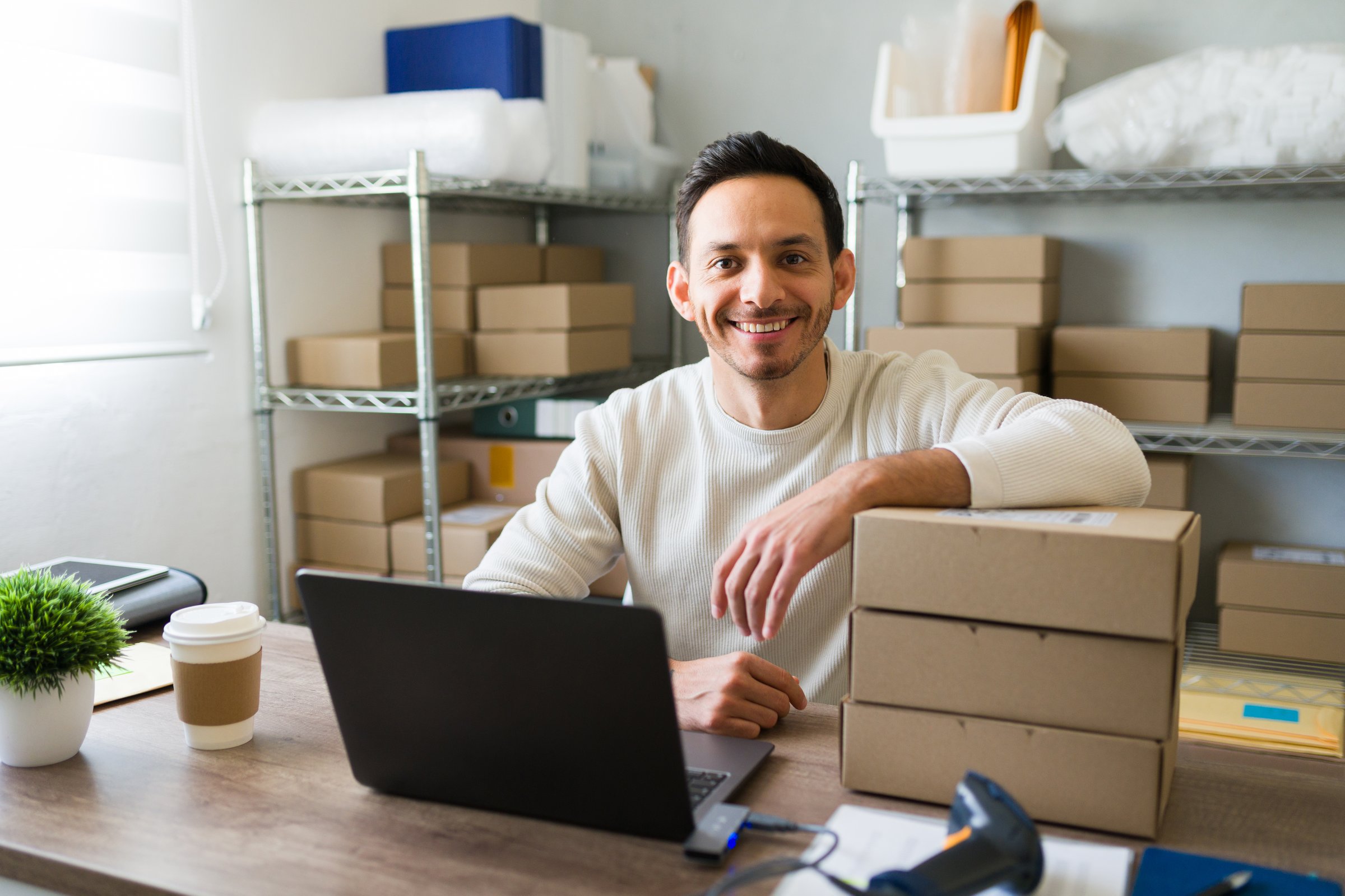 Happy latin business owner working on shipping packages from his home office using a laptop and a barcode scanner