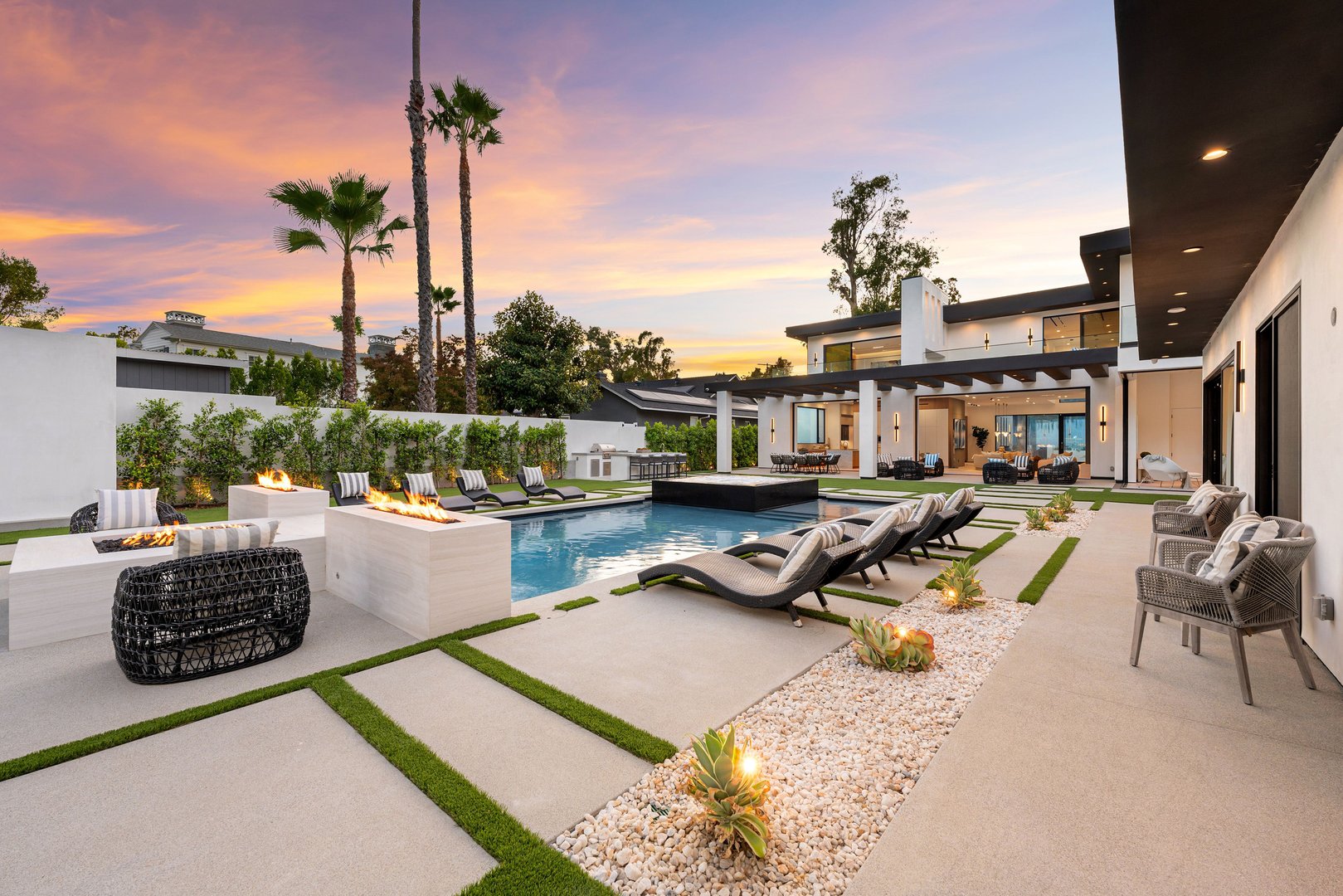 Luxurious modern backyard with pool, fire pits, and lounge chairs at sunset, featuring palm trees and elegant landscaping