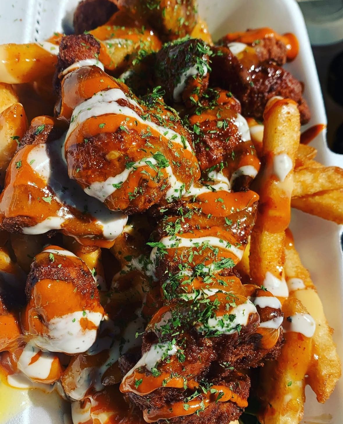 Crispy chicken bites and fries covered in orange sauce and ranch, garnished with herbs in a takeout container.