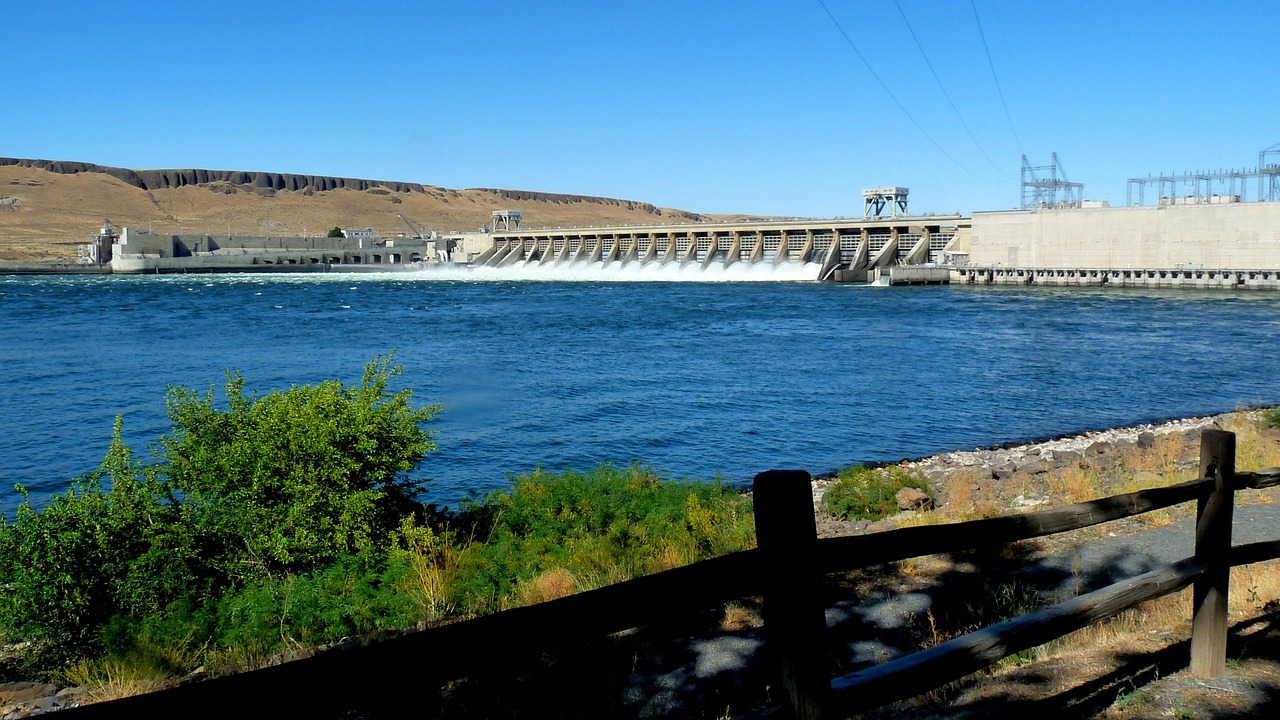 Columbia River dam with water flowing over spillway, surrounded by arid landscape and a wooden fence in the foreground.
