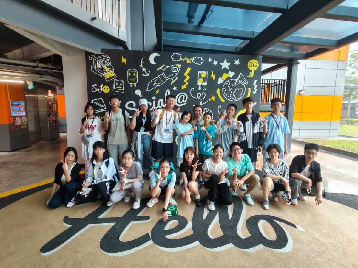 Group of young people posing in front of a colorful wall mural with Hello on the floor. Indoor casual setting.