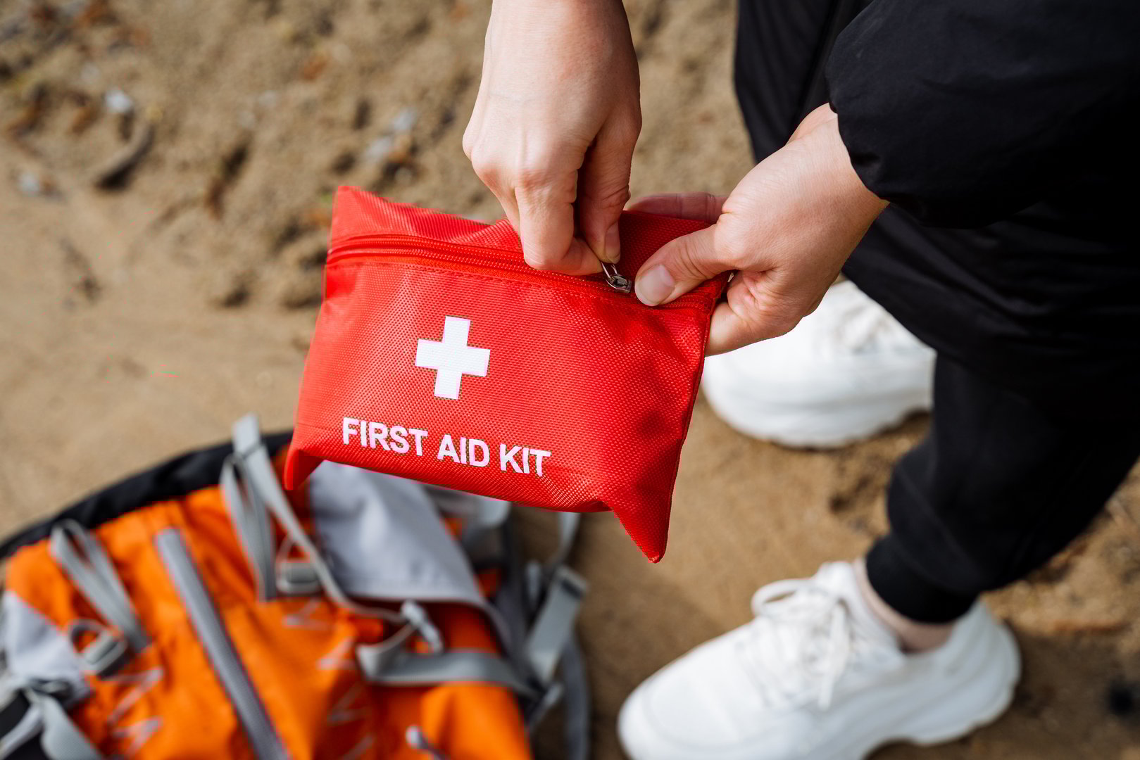 Hands carefully preparing a first aid kit on a sandy beach, emphasizing the importance of outdoor safety and emergency preparedness. Backpack in the background for travel health and protection