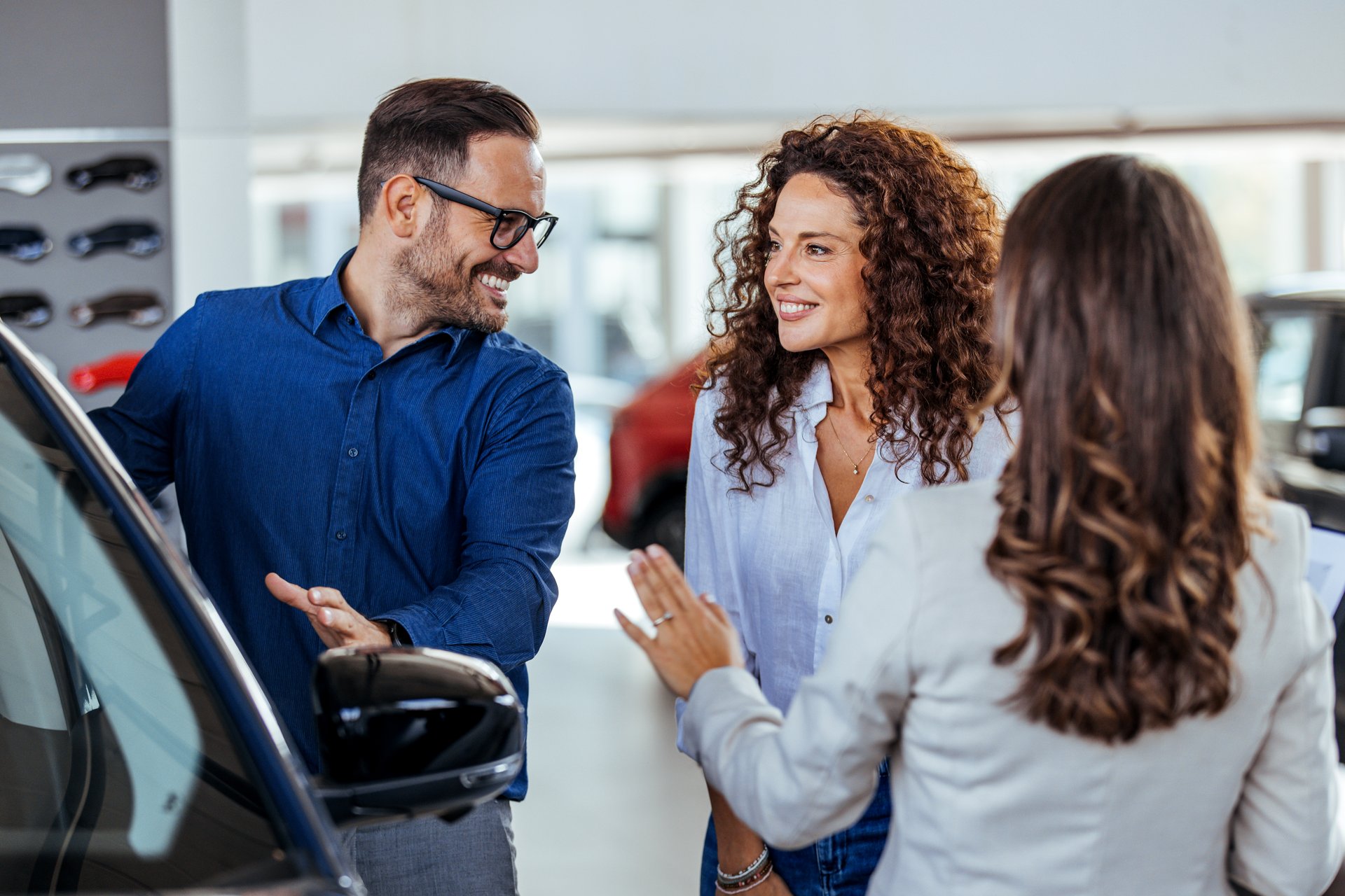 A car salesman is showing new cars to a couple. Car sales business. Manager talking to couple, showing them new auto at dealership shop. Young family selecting vehicle, looking at modern automobile at showroom