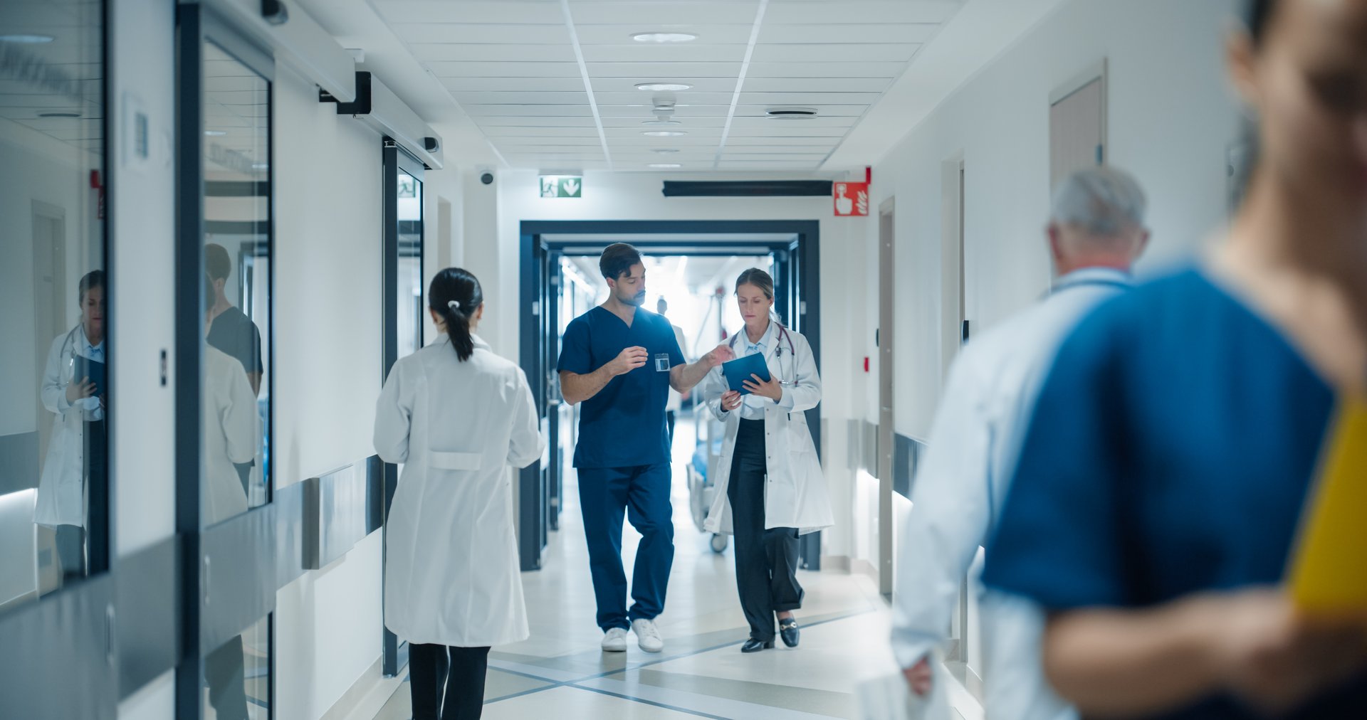 Medical Staff Navigating a Busy Hospital Hallway. Caucasian Male Doctor and Female Nurse Having a Discussion About an Upcoming Surgery and Medical Procedures as They Walk in Corridor with Other People