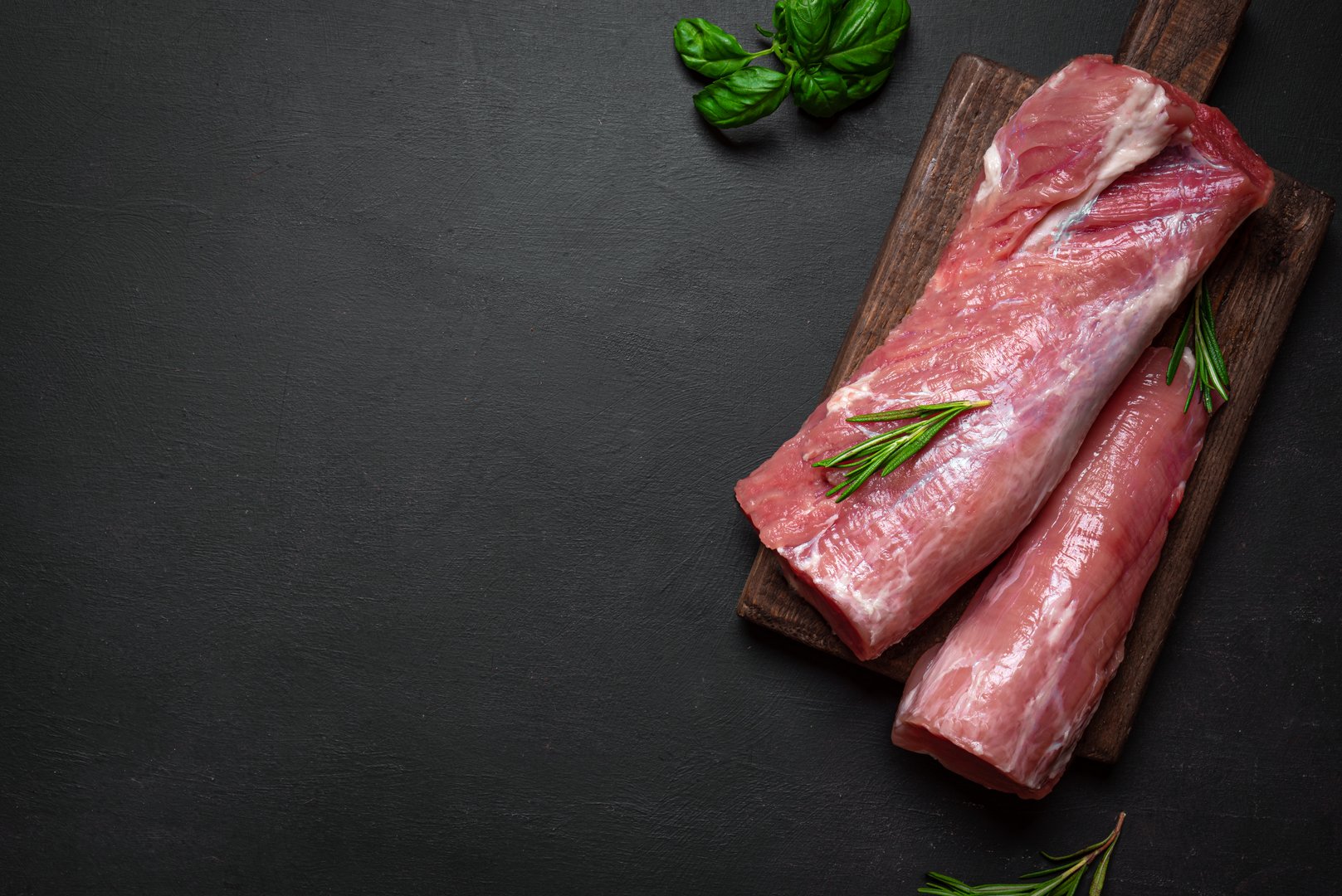 Uncooked pork tenderloin meat ready to cook on wooden background with rosemary, top view, copy space. Fresh juicy meat ready to cook, food concept.